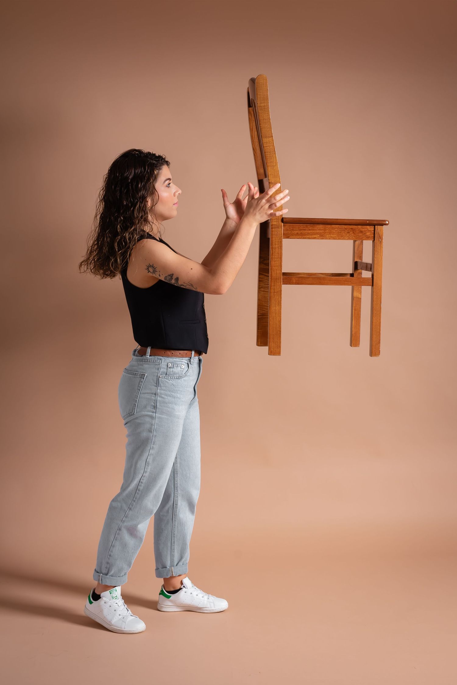 Person in black top and jeans playfully holding up a wooden chair against beige background.