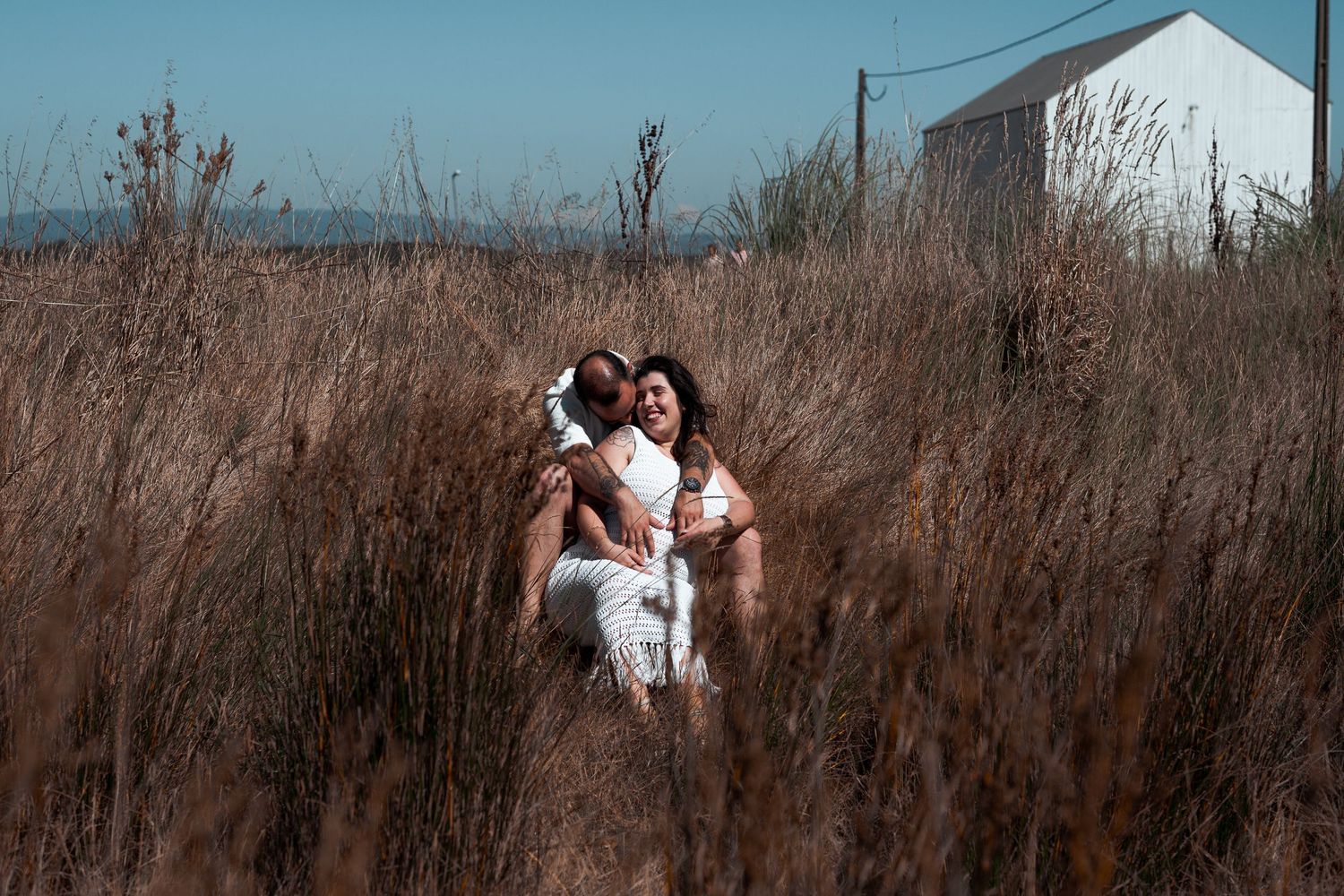 Couple in white outfits standing among tall dry grass with white building in background.