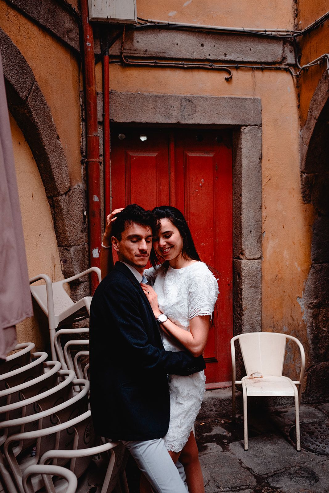 Couple embracing near a red door with plastic chairs in an outdoor courtyard setting.