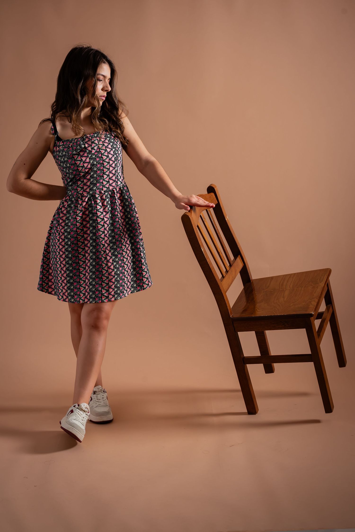 Model in patterned dress poses playfully with wooden chair against peach colored background.