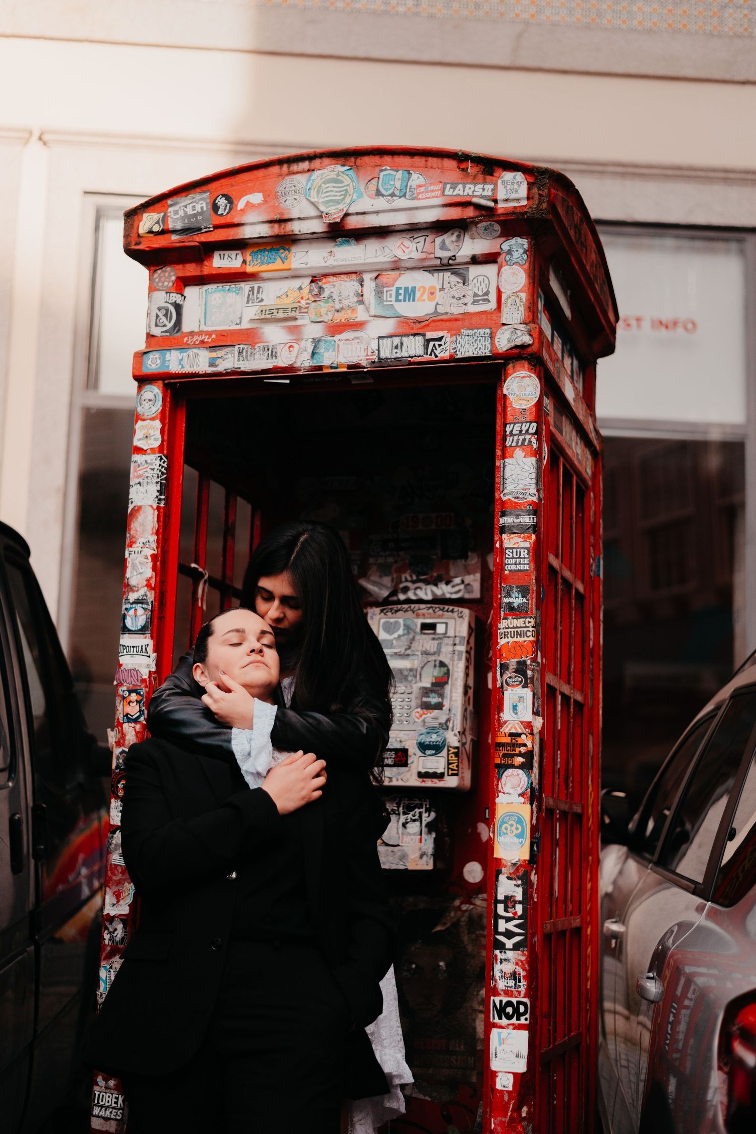 A couple embraces tenderly beside a red British phone booth covered in colorful stickers on a city street.