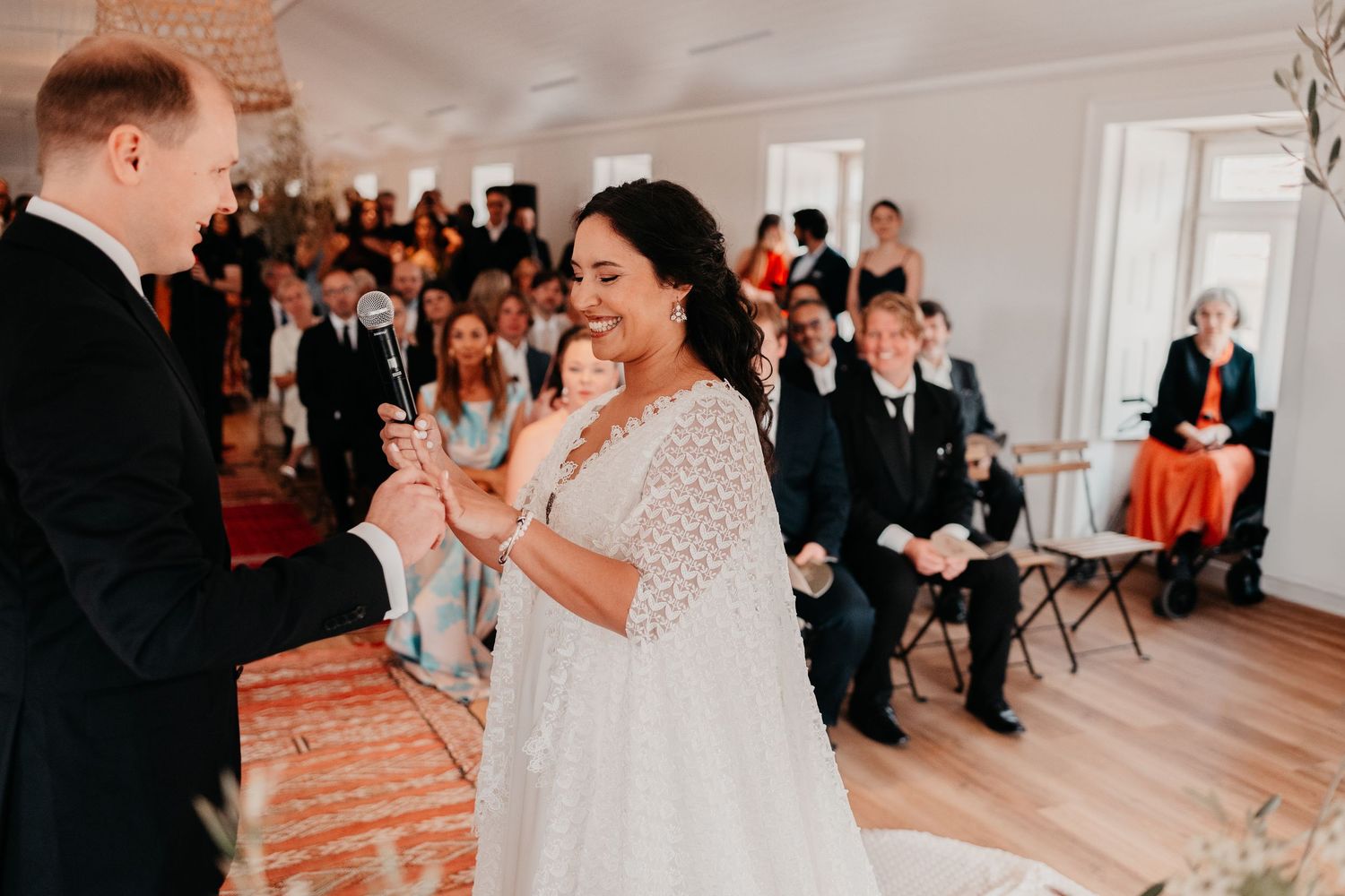 Bride in white lace gown smiling during indoor wedding ceremony ring exchange with guests seated in background.