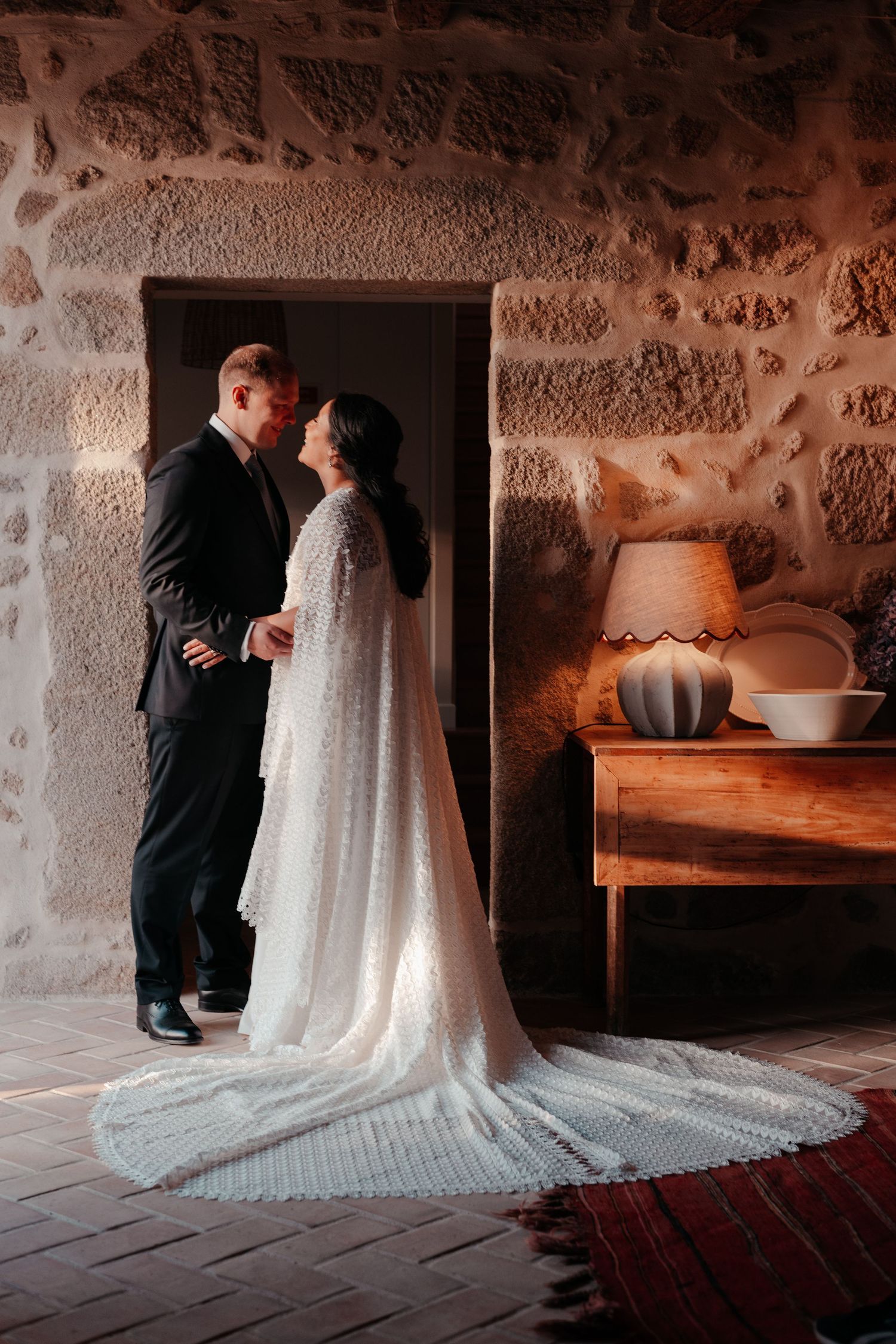 Bride in white lace cape gown and groom in black suit share romantic moment in rustic stone venue.