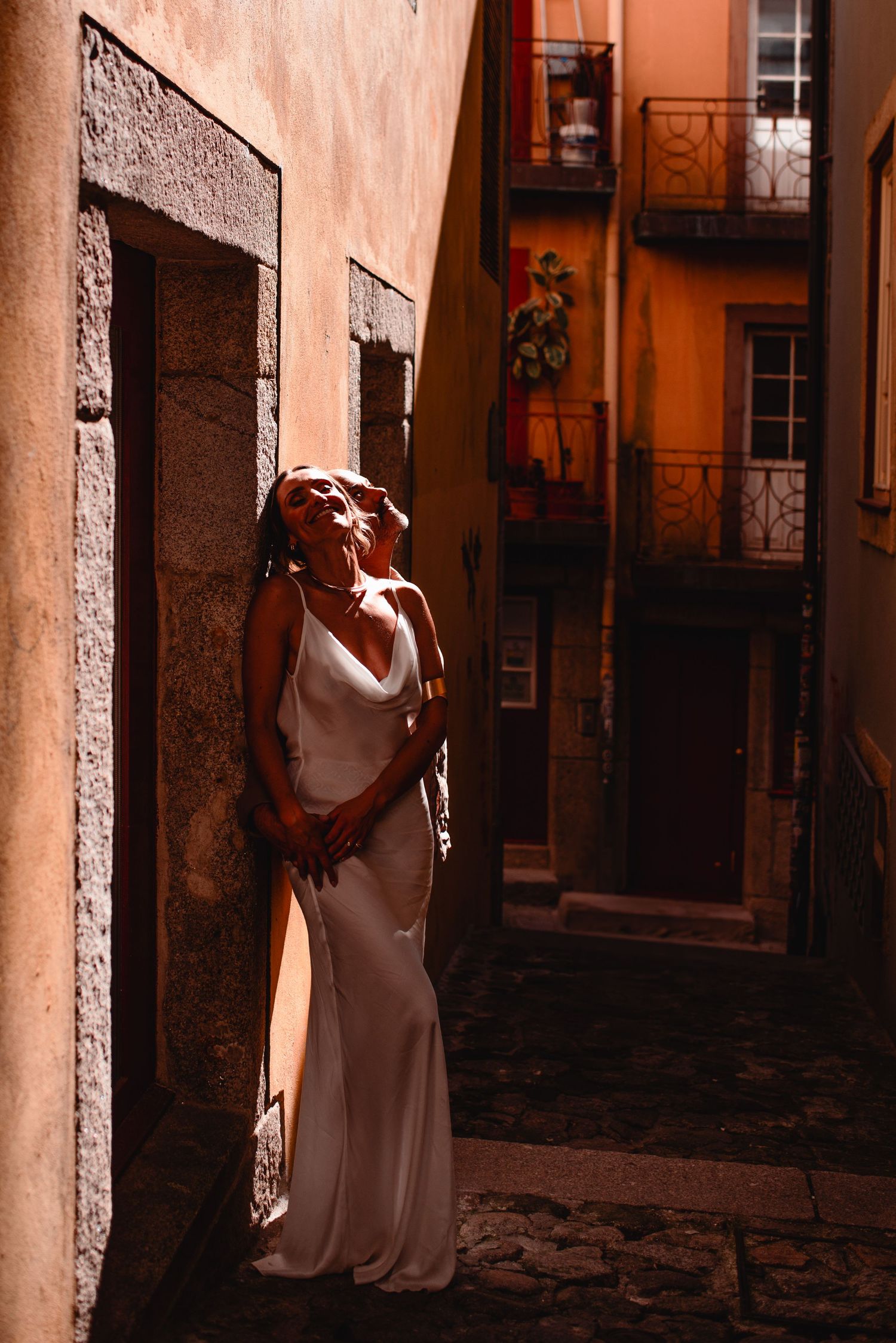 A person in a white dress poses dramatically against terracotta walls in an urban setting.