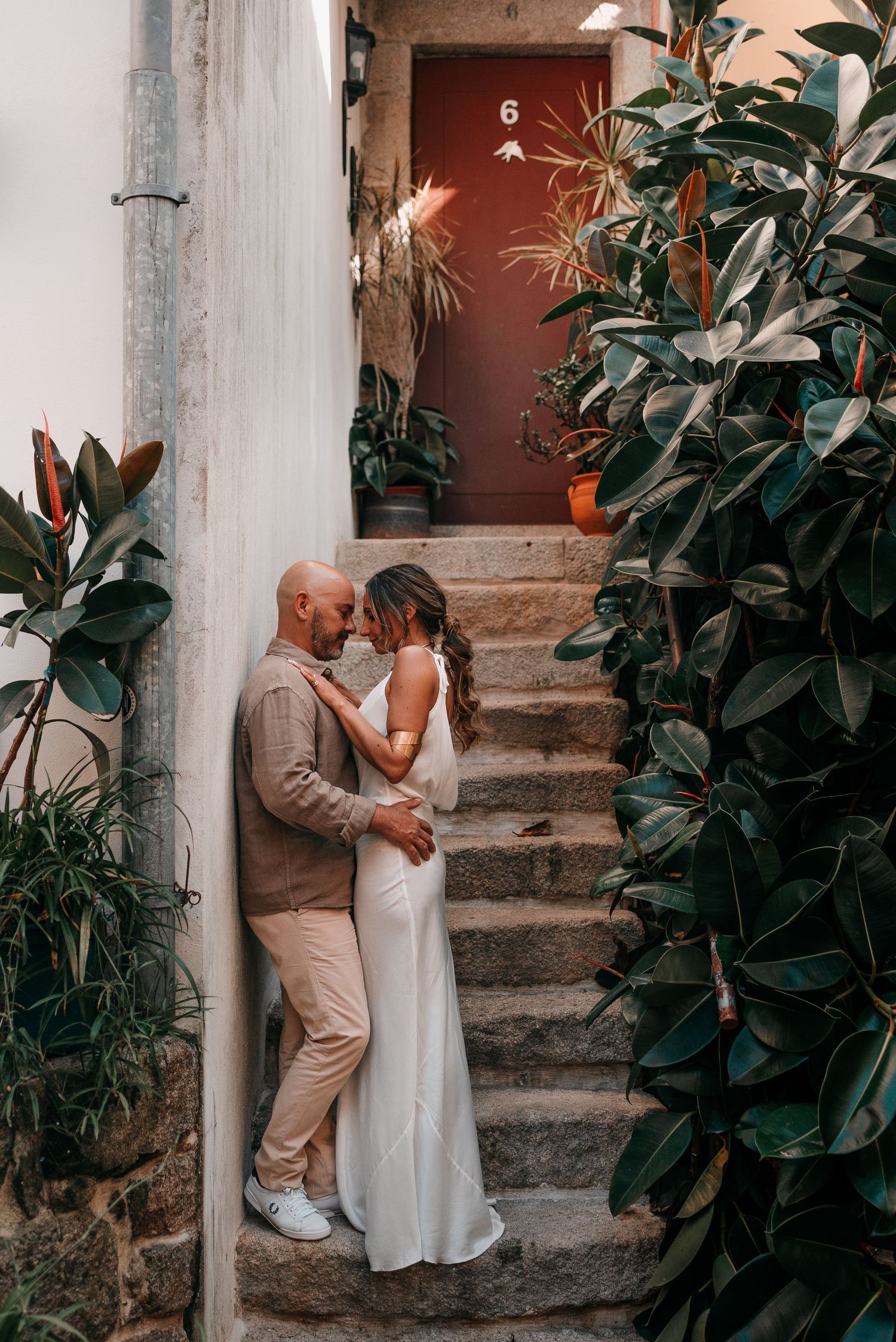 Couple embracing on stone steps surrounded by lush plants near a red door, bride in white gown.