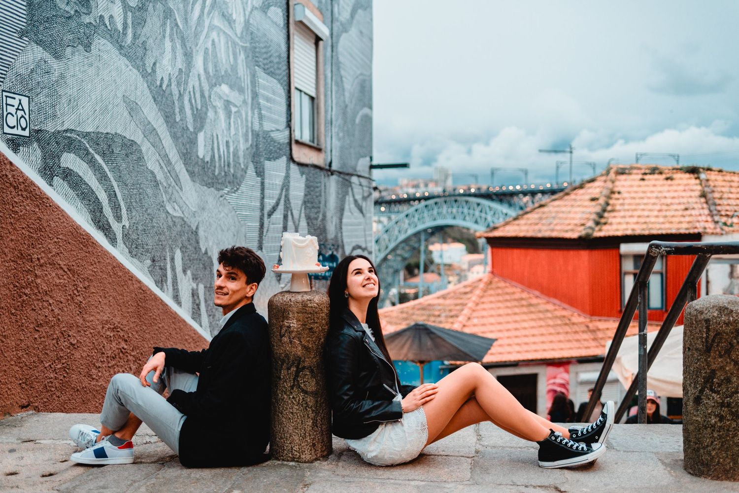 A young couple sits back-to-back on a stone ledge with colorful rooftops and street art mural behind them.