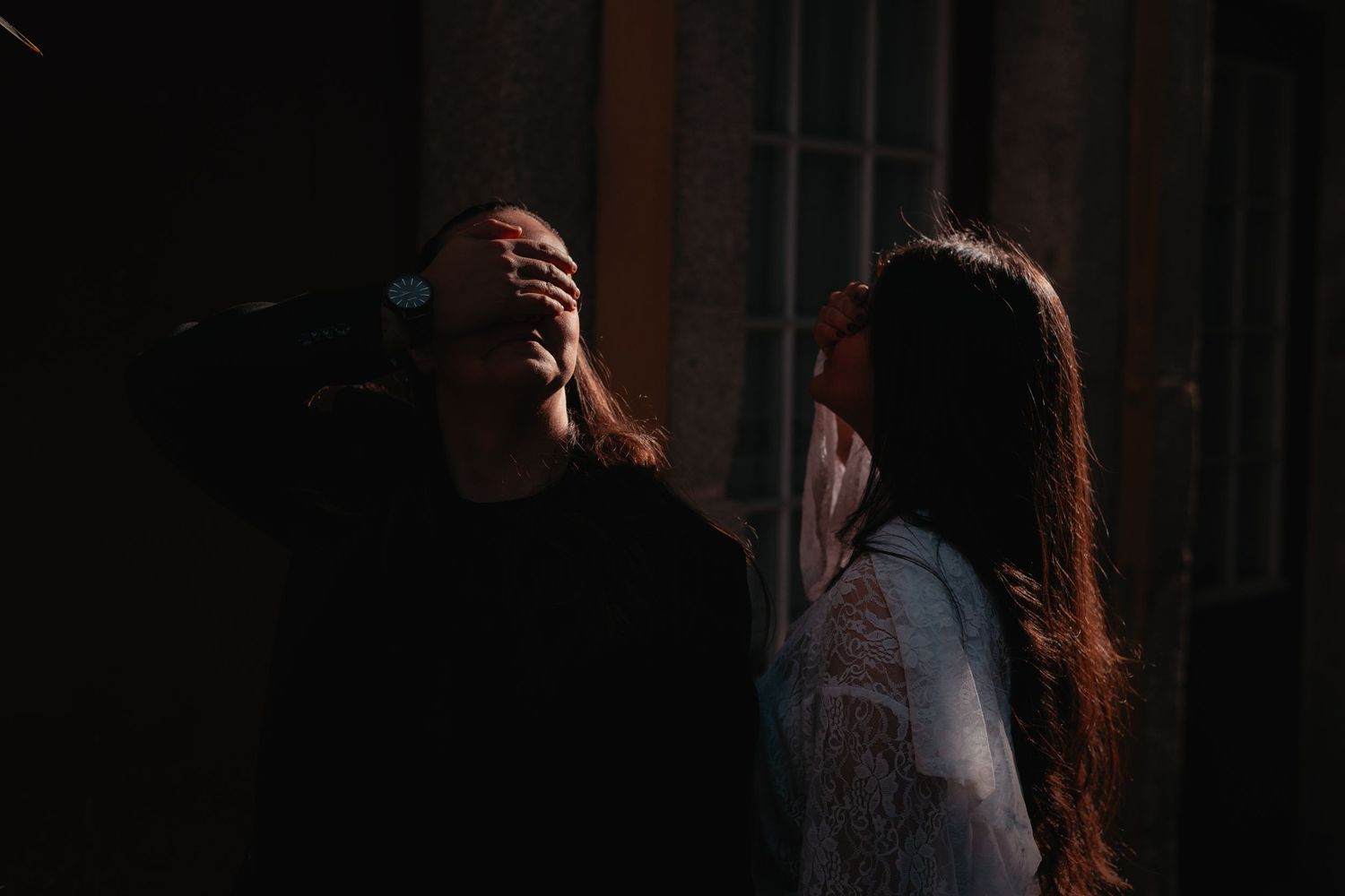 Two women in dramatic lighting, one covering her face with her hand while the other looks on in a dark setting.