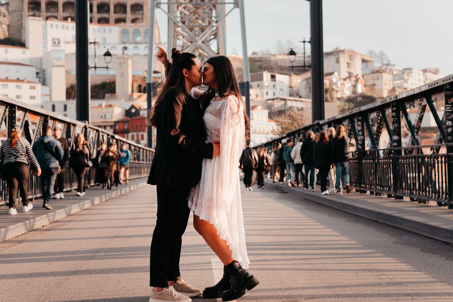 Couple kissing on a bridge in Porto, Portugal, with tourists and the Dom Luis I Bridge in the background.