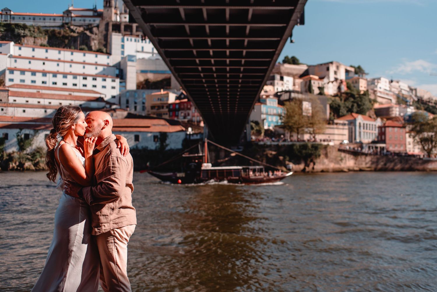 Couple poses along riverfront with historic Porto buildings and bridge in background.