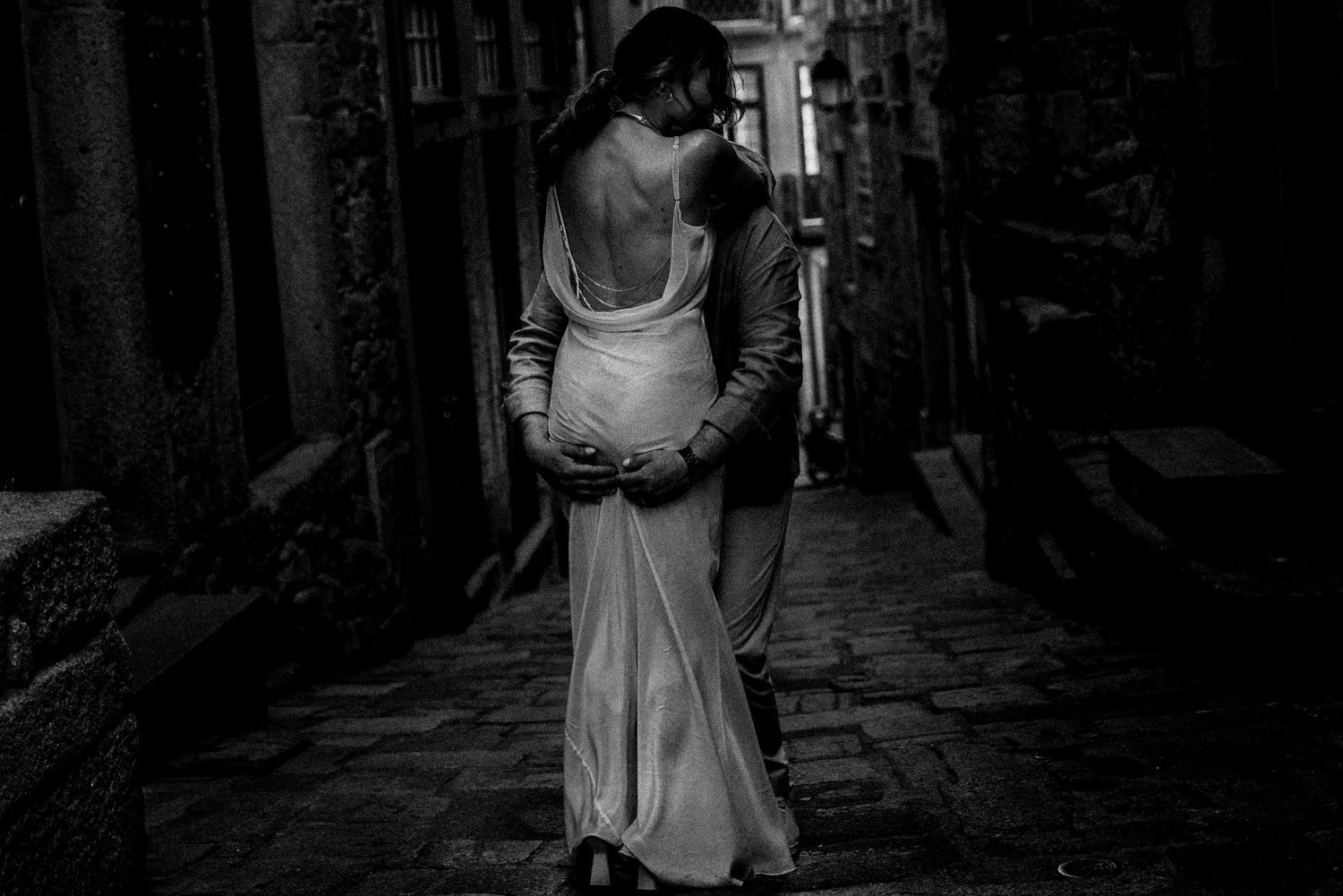 A backlit silhouette in a flowing dress walks through a narrow cobblestone alley.