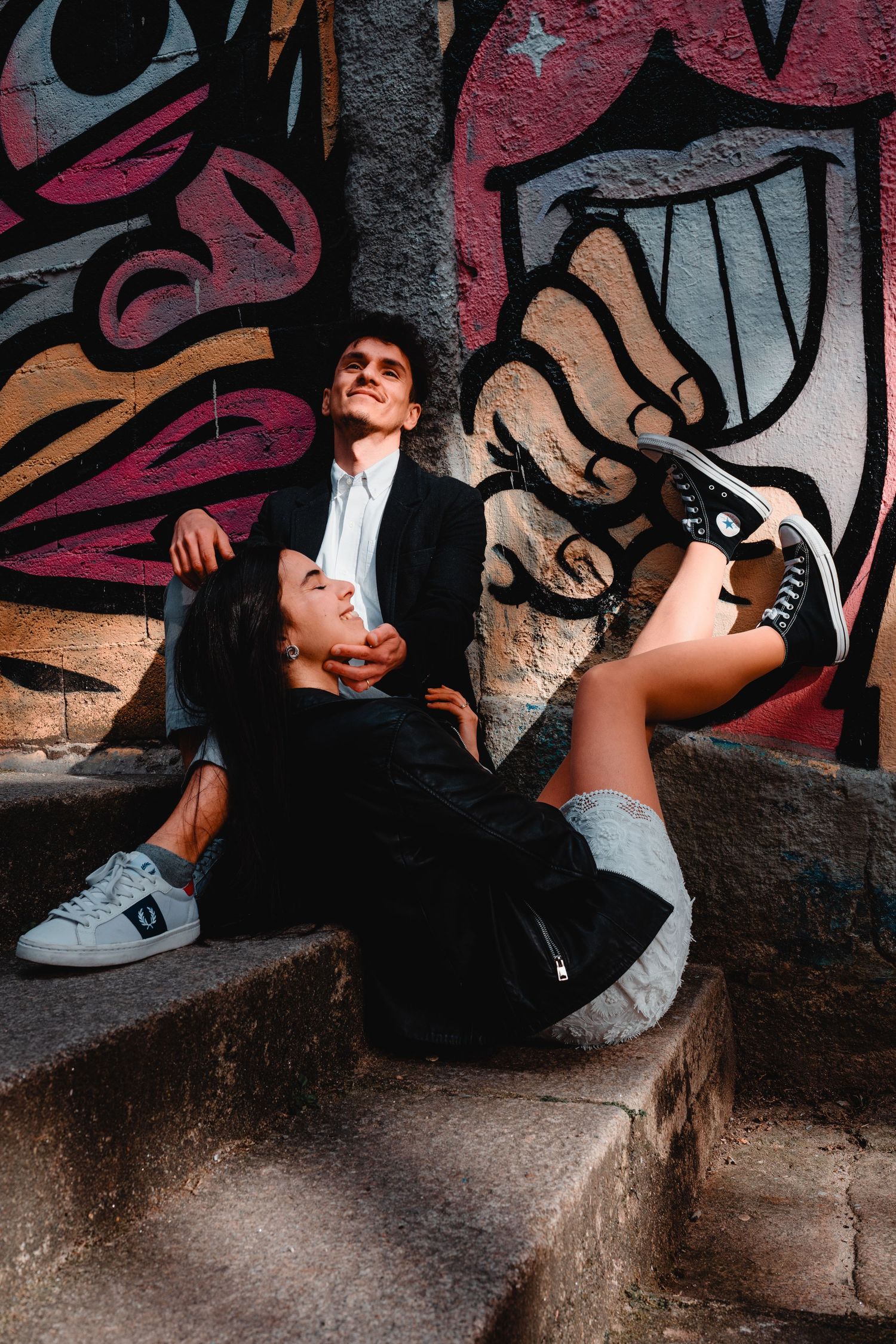 Couple sitting on stone steps against colorful graffiti wall, woman leaning back smiling up at man in black suit.