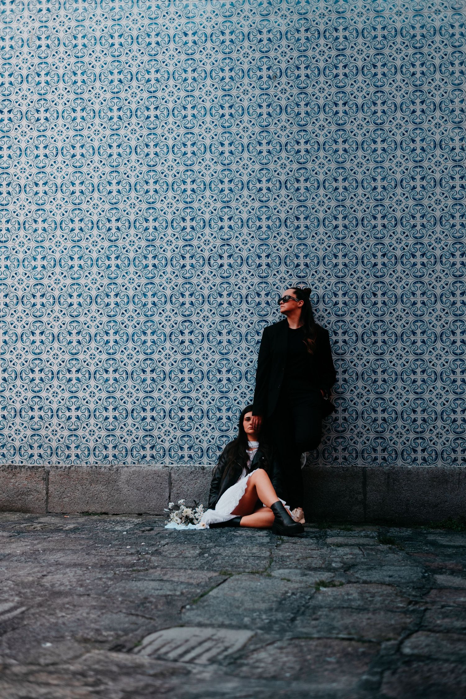 Couple posing against a blue decorative tiled wall, one standing and one sitting on cobblestone ground.