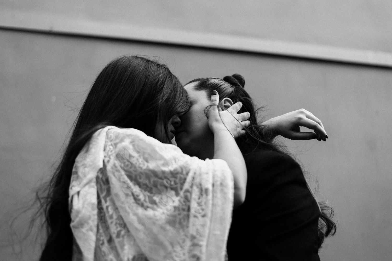 Two women share an intimate moment, one covering her face, in a black and white street photography shot.