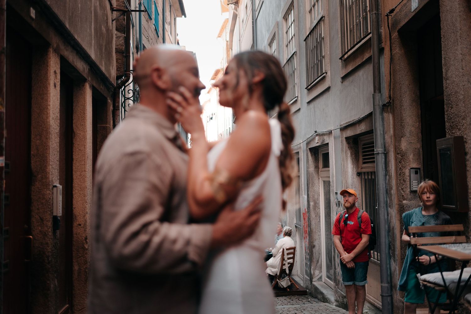 Couple sharing romantic kiss in narrow European alley, onlookers in background, wedding attire.