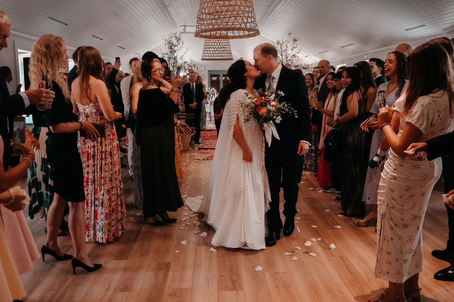 Bride and groom share a kiss at their wedding reception while guests toss flower petals in a bright, elegant venue.
