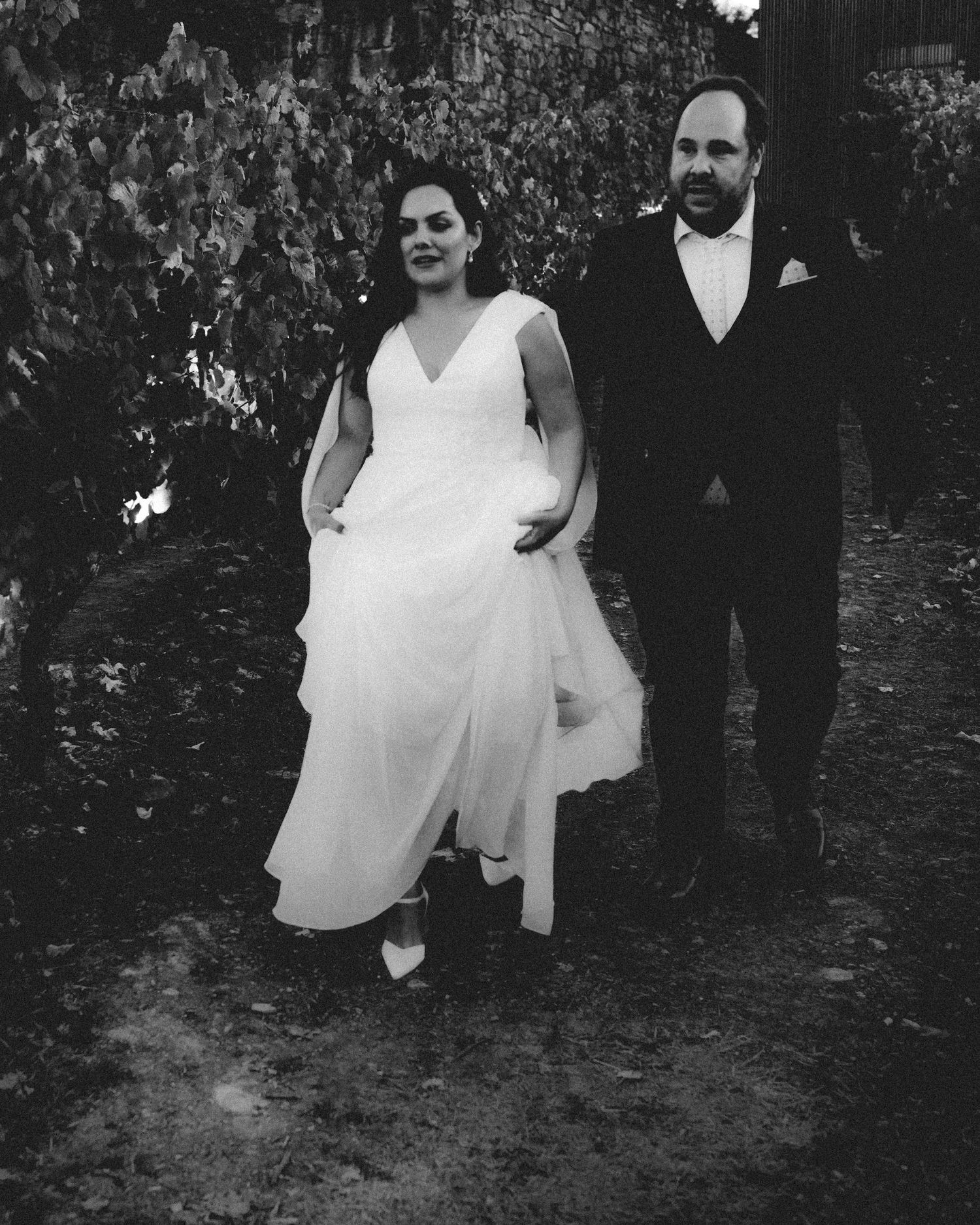 Black and white photo of a bride in white gown and groom in dark suit walking together outdoors.