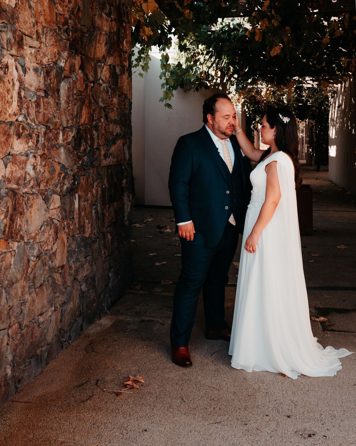 A bride in a white cape gown and groom in navy suit share a tender moment outdoors near a stone wall.