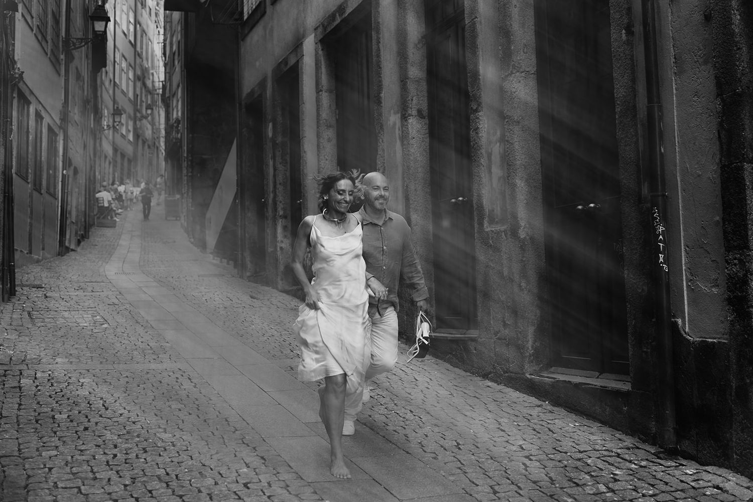 Couple walking through a cobblestone alley in dramatic black and white with sunbeams streaming through.