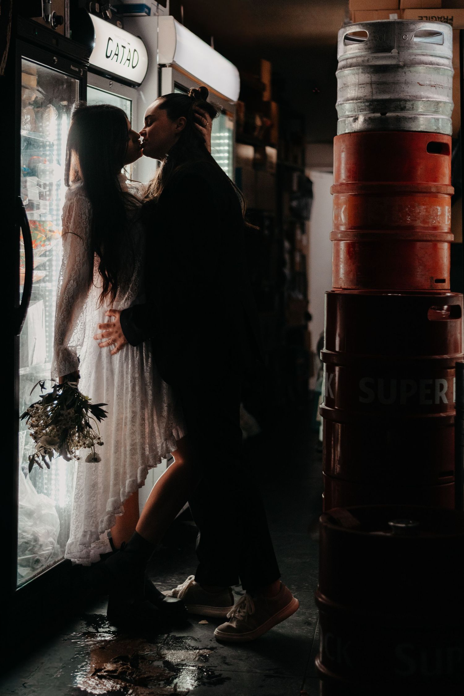 Couple sharing a romantic kiss in a dimly lit bar, bride in white dress holding bouquet beside stacked beer kegs.