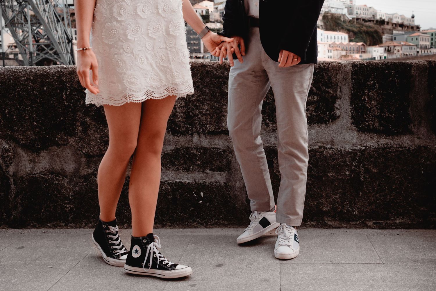 Couple holding hands in casual attire, woman in white dress and Converse sneakers, man in gray pants, standing on stone pavement.
