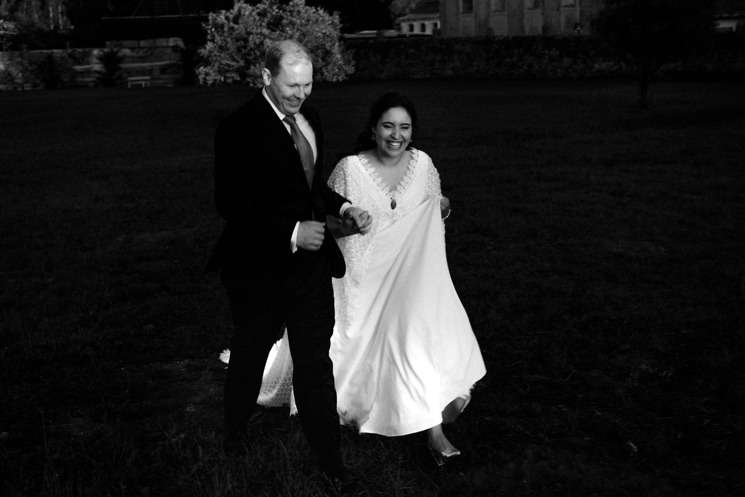 Black and white photo of a couple in formal attire walking and laughing together on a lawn.