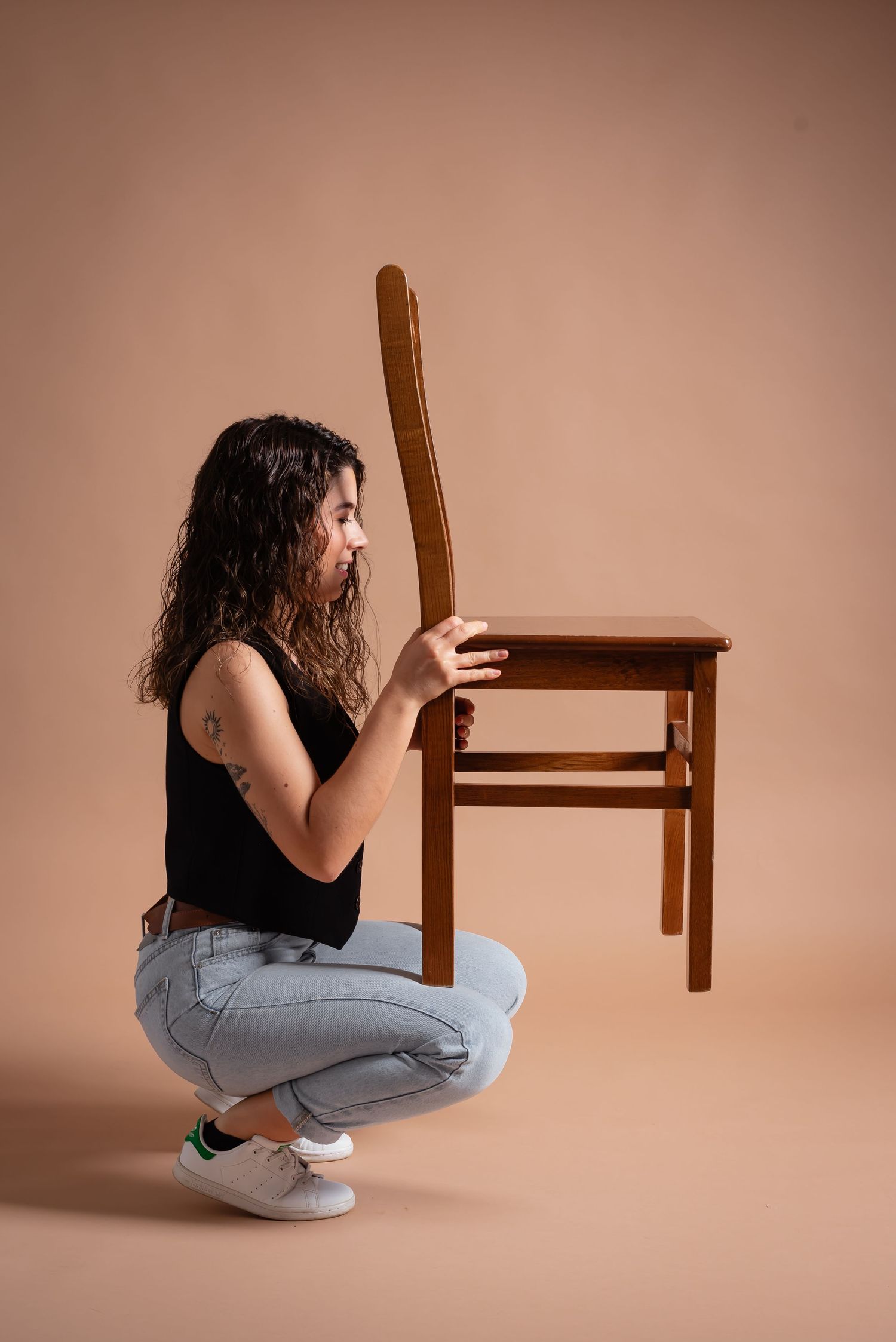 Figure examining wooden chair while crouching in casual attire against peach backdrop.