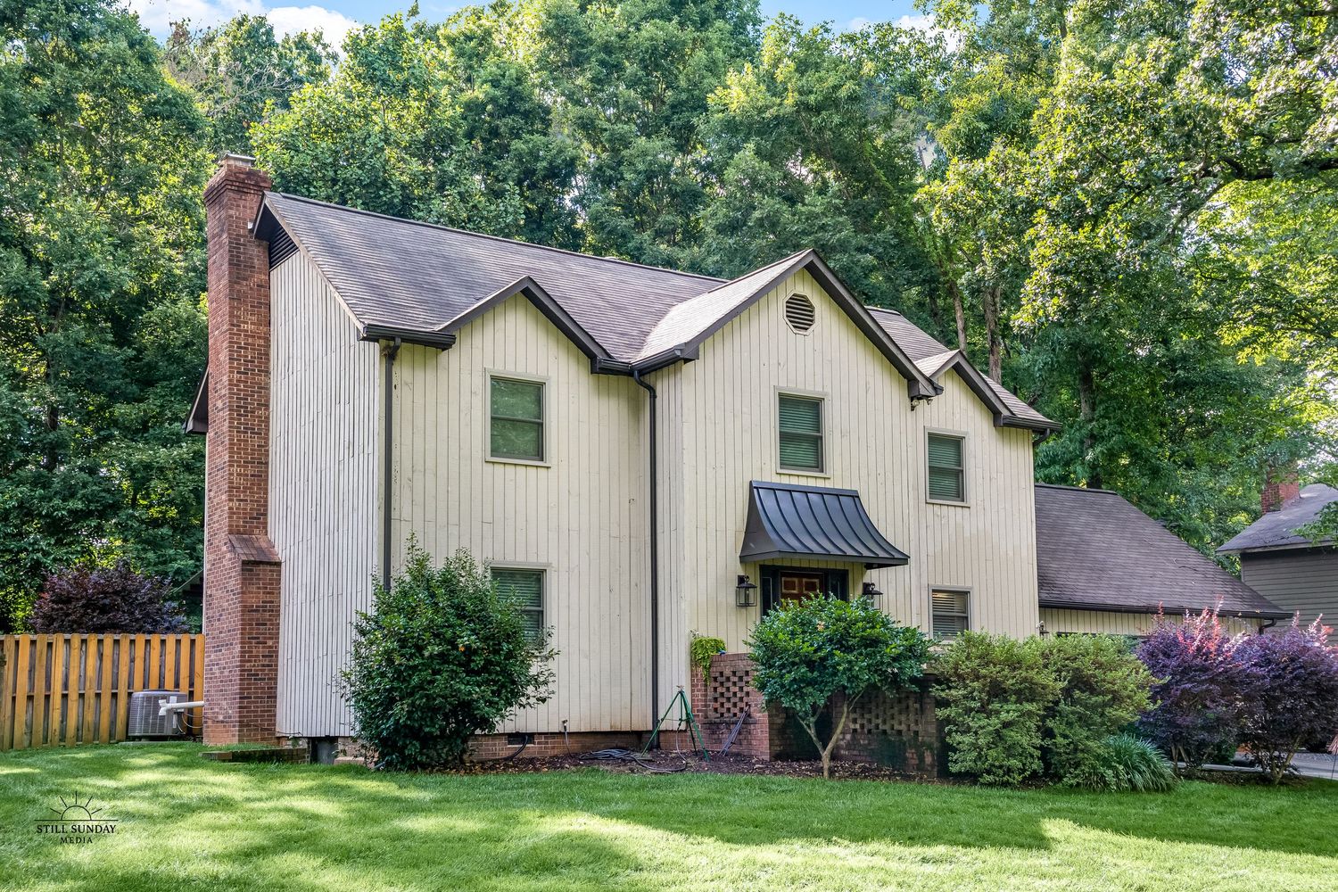 Front exterior real estate photo of a beige two-story home with a brick chimney and lush landscaping