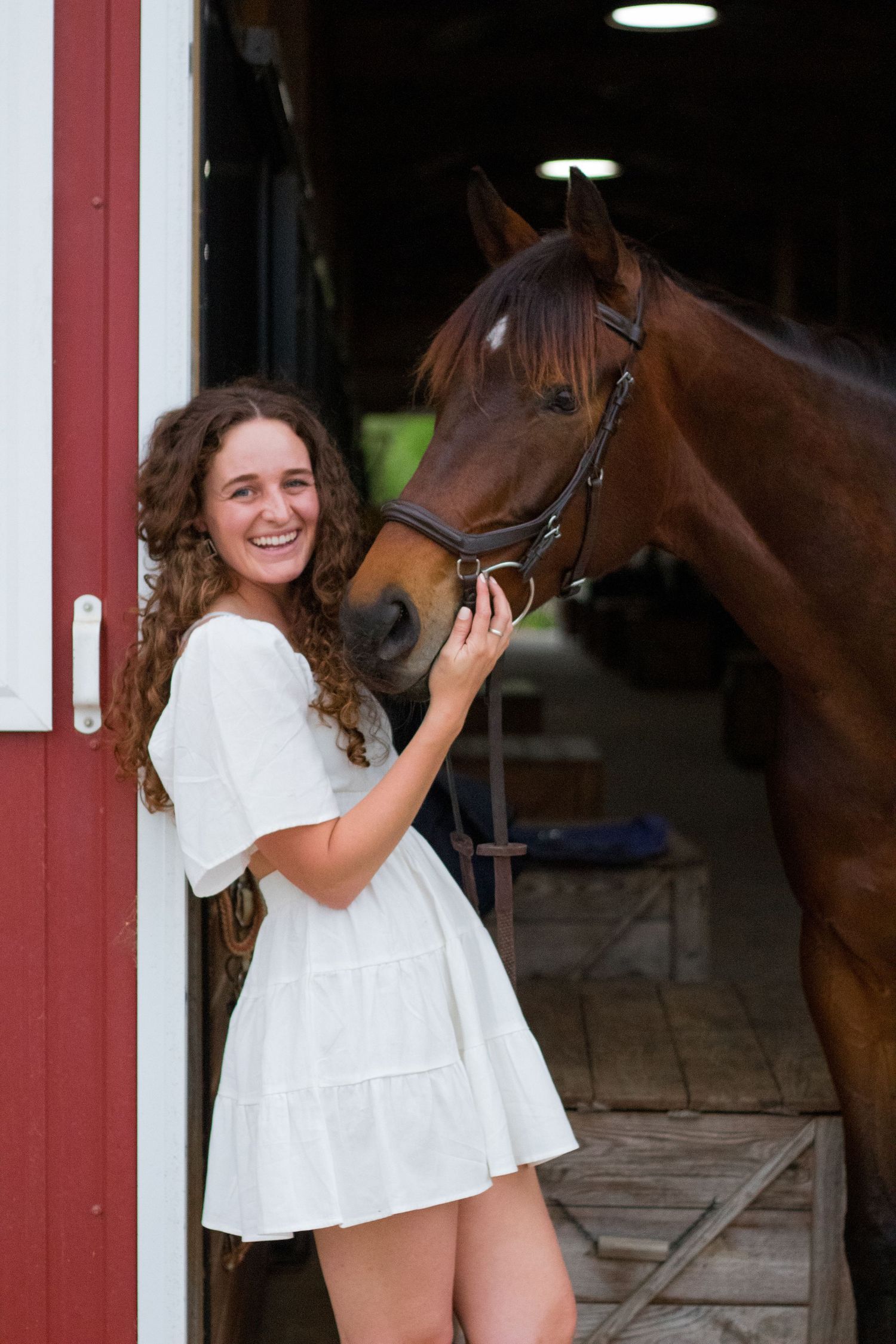 Woman in white dress petting her horse outside a red barn for an equine portrait session in North Carolina