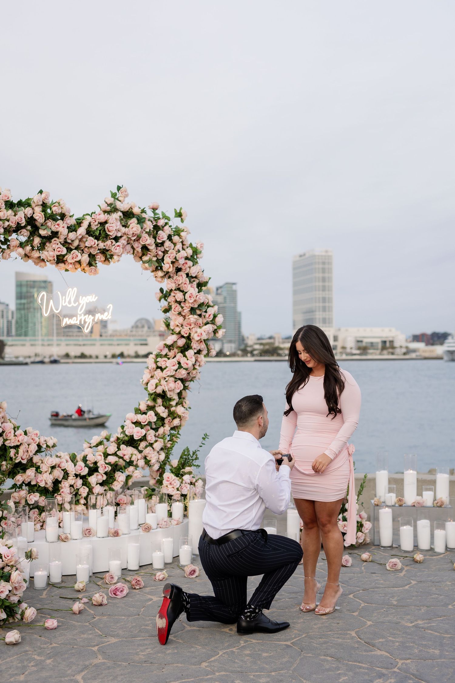 Romantic proposal setup by the waterfront with floral arch and candles as couple shares intimate moment.