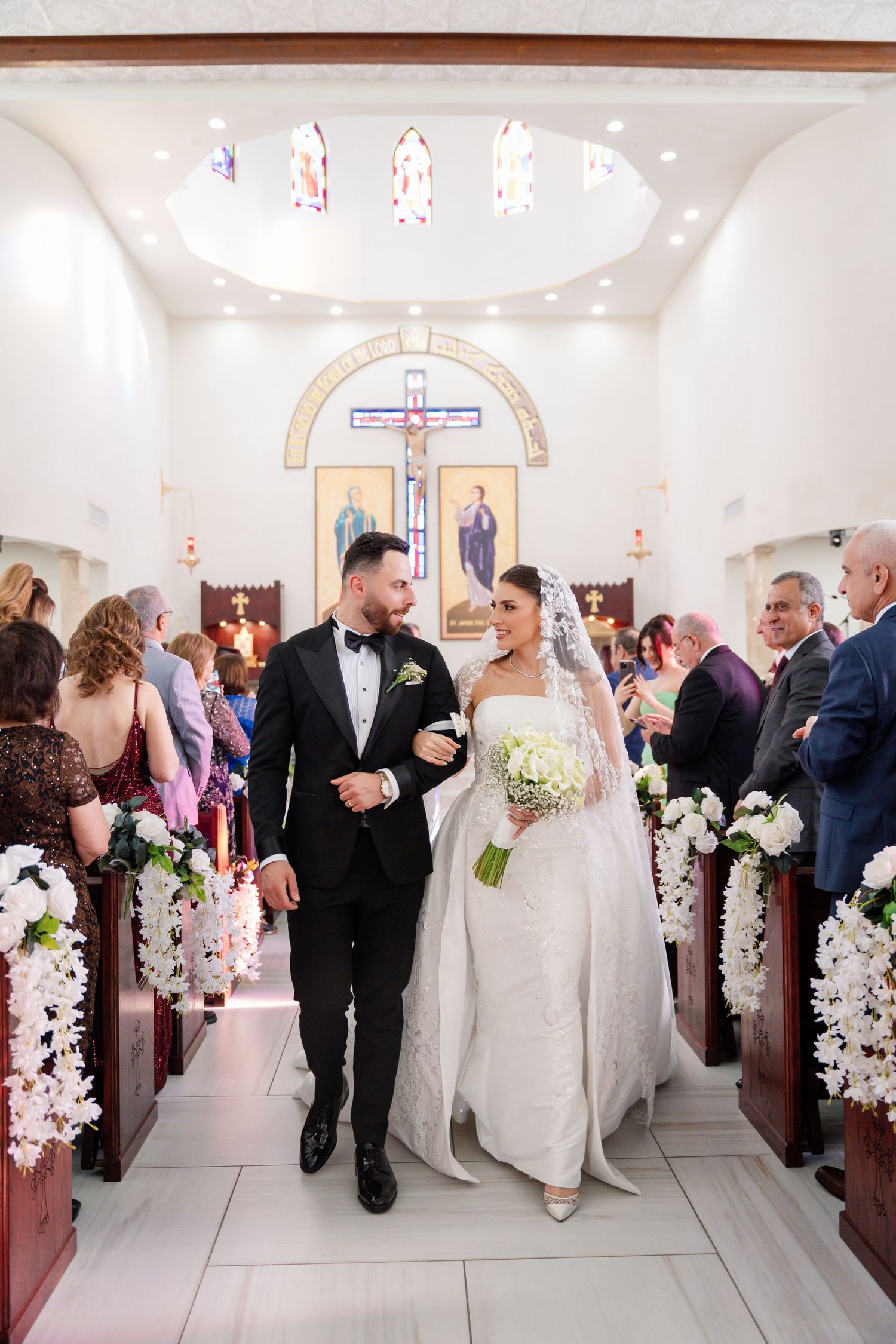 Wedding procession in a modern church with white floral decorations along the aisle.