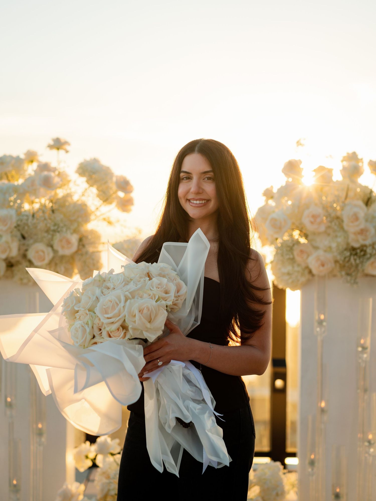 Beautiful white floral arrangements with roses line an outdoor venue at sunset.