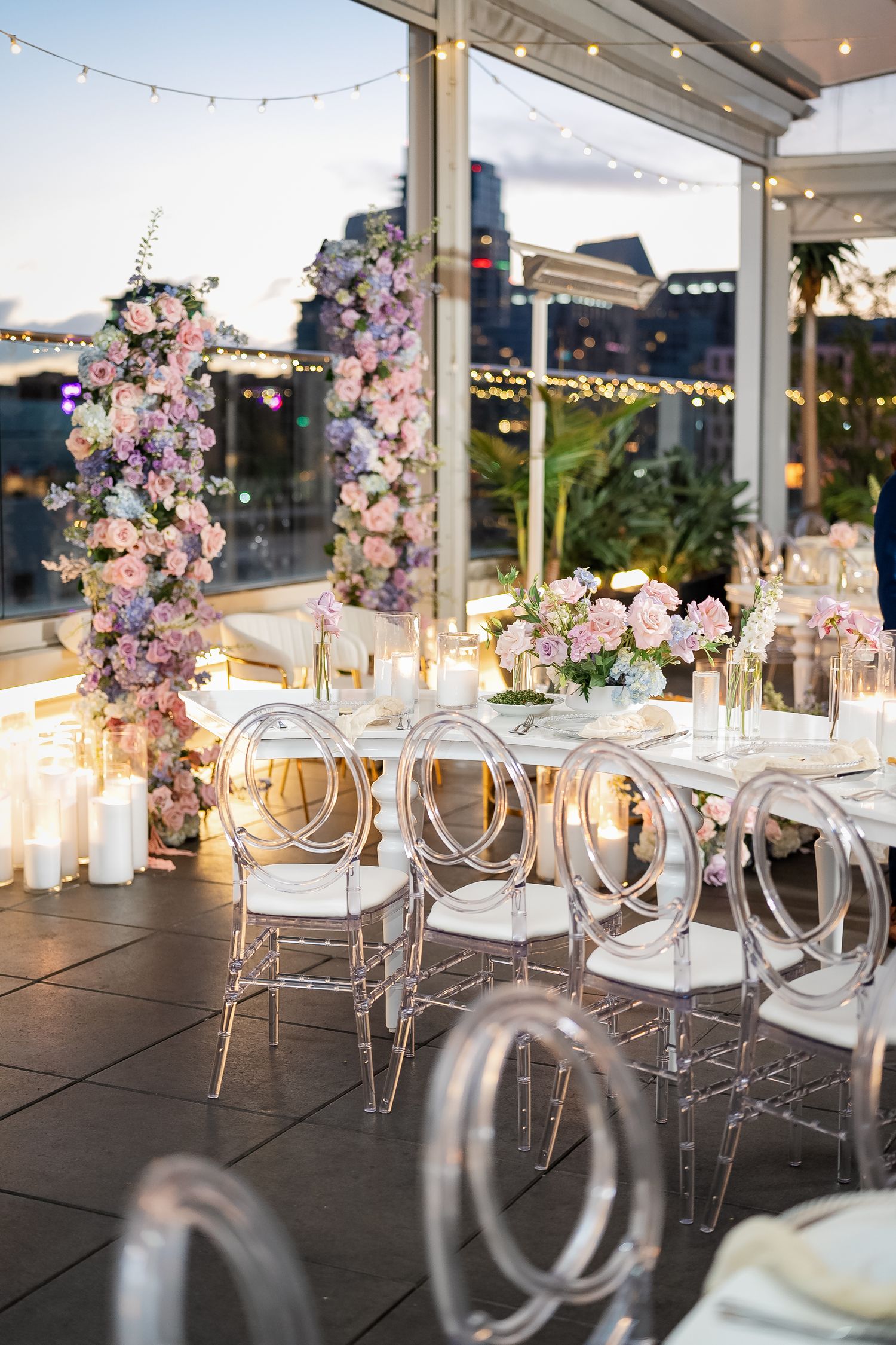 Modern wedding reception setup with ghost chairs and floral arrangements under string lights.