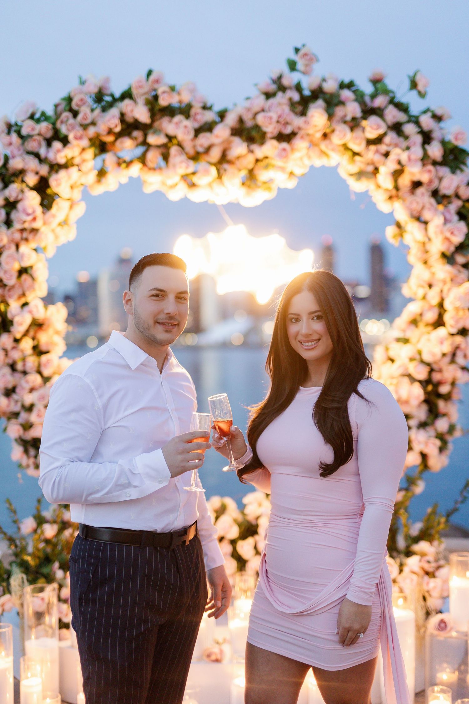 Newly engaged couple toasting with champagne under lit floral arch by waterfront.