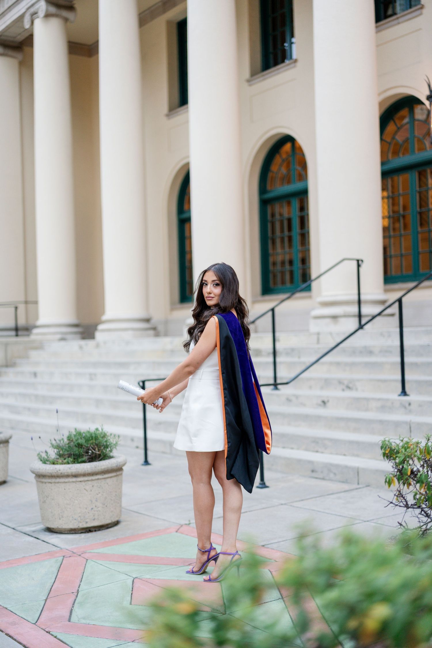 A graduate in white dress poses playfully on classical stone steps with white columns.