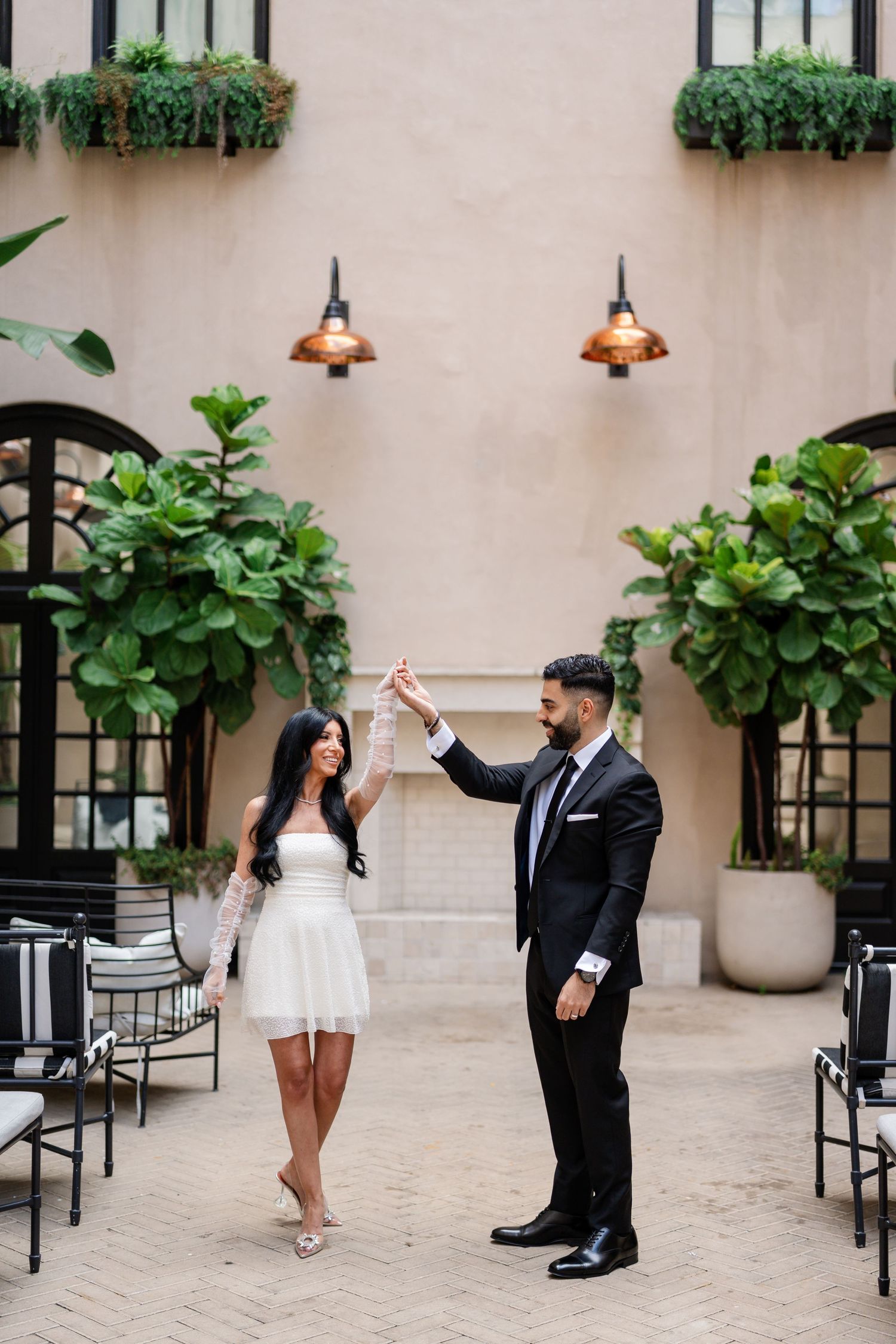 Couple in formal attire posing playfully in courtyard with arched windows and tropical plants.