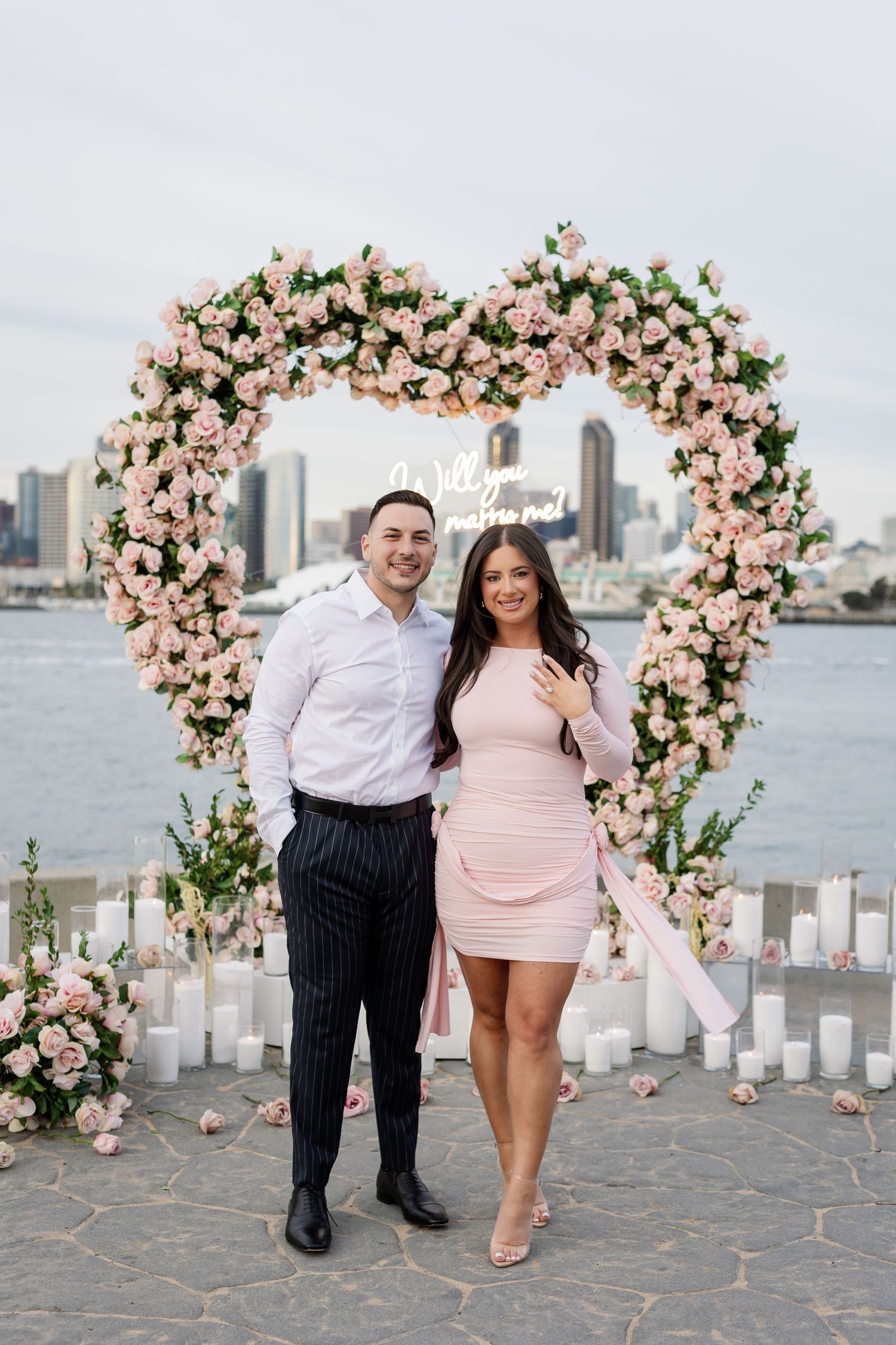 Couple poses by heart-shaped floral arch with pink roses overlooking city skyline.