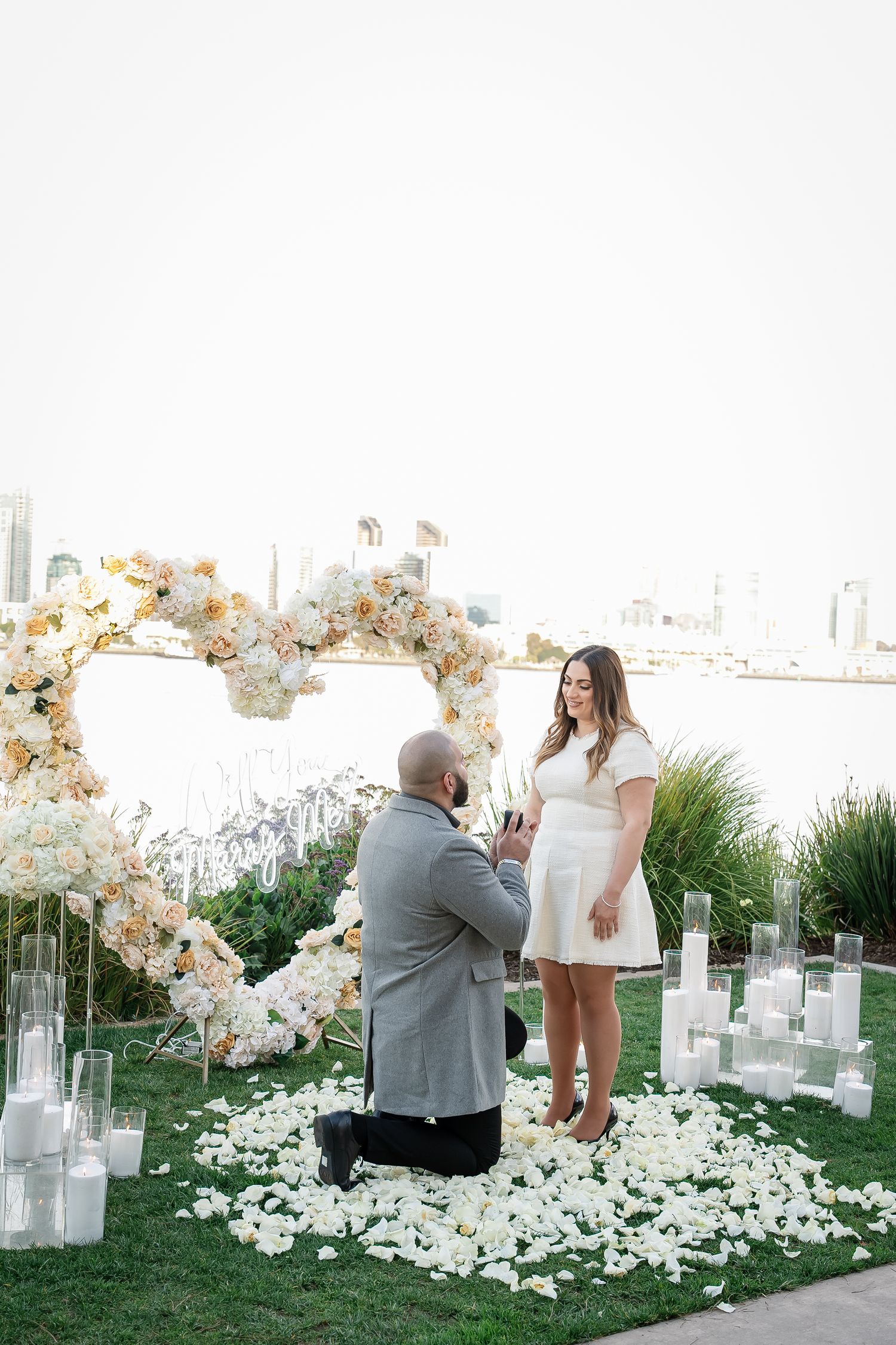 Outdoor proposal scene with white orchid arch and scattered rose petals on grassy setting.