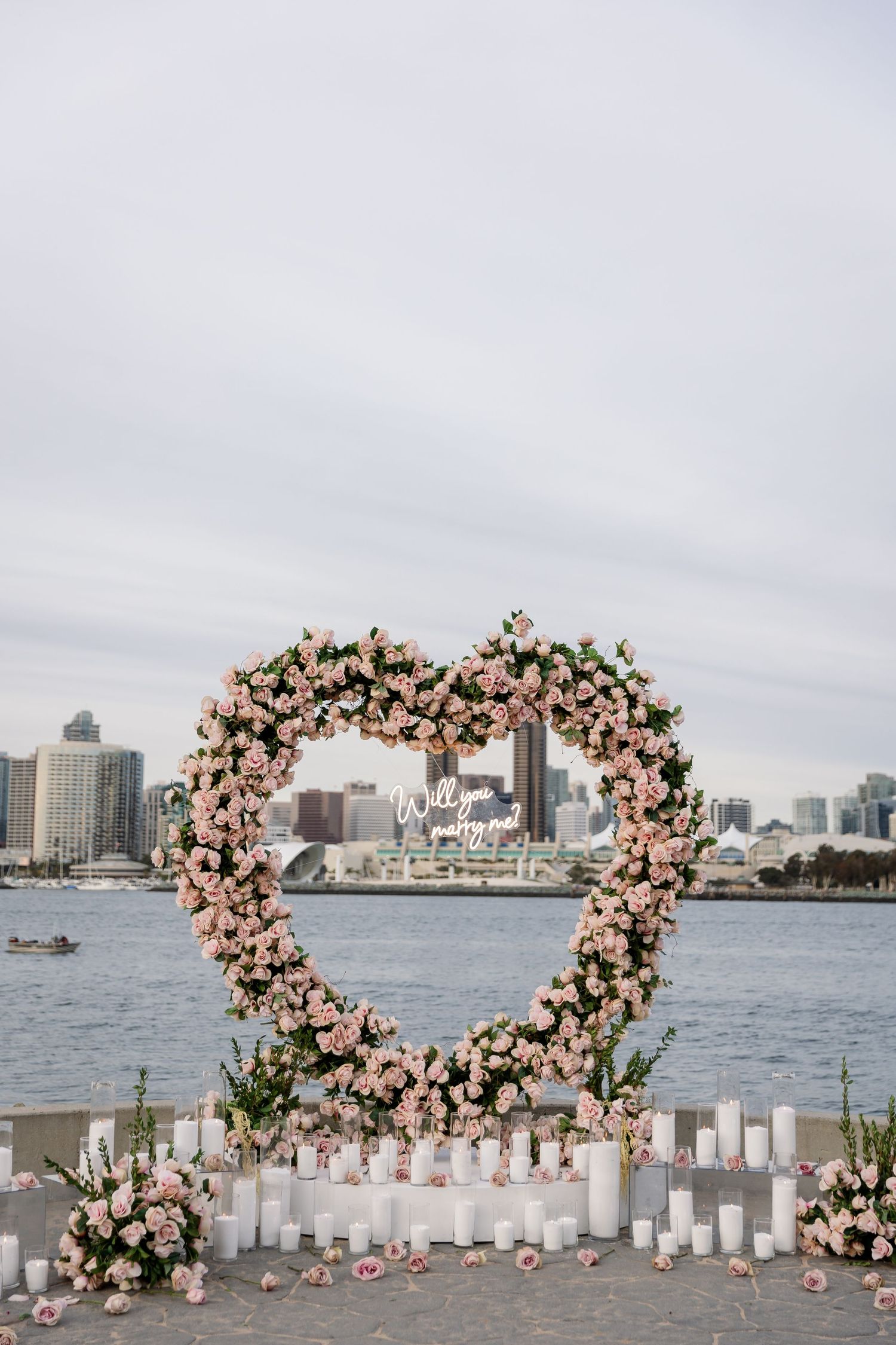Pink floral heart arch with white candles frames waterfront cityscape view.