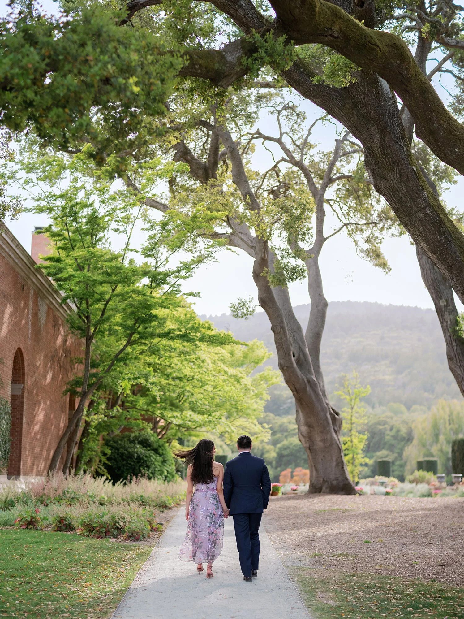 A couple strolls along a tree-lined path with mountains visible in the distance.