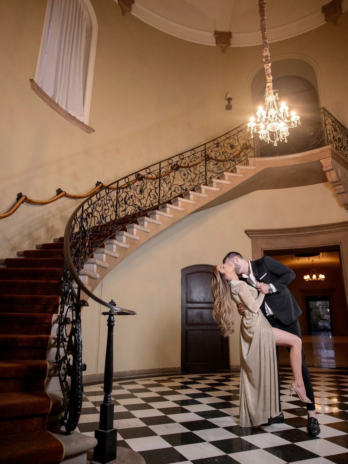 Black and white checkered floor with ornate staircase and chandelier in a grand ballroom setting.