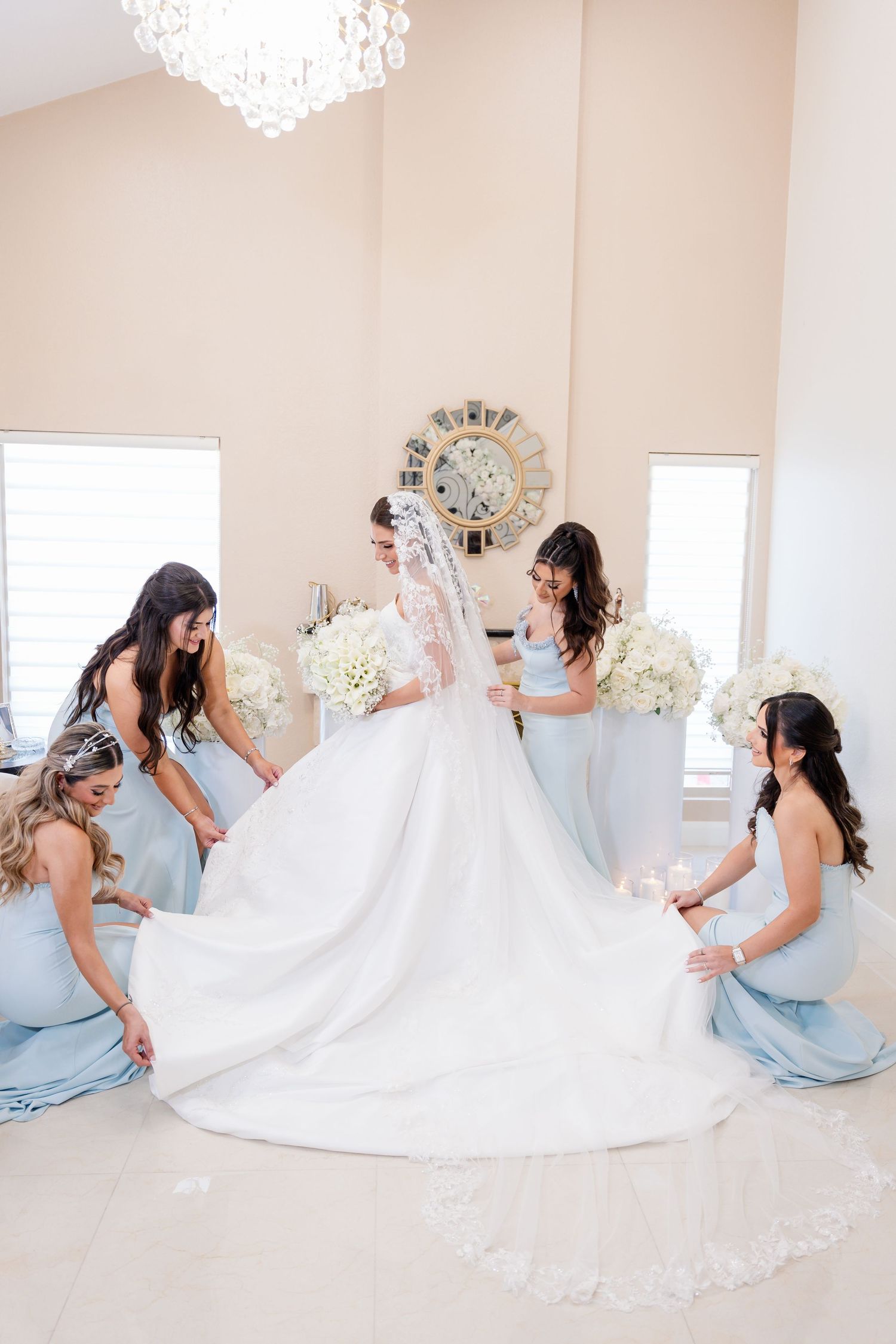 Bridesmaids in light blue dresses help arrange a wedding gown's train in a bright white room.