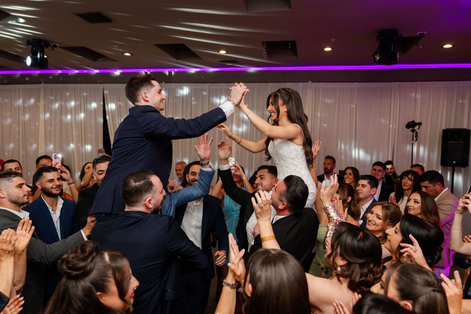 Bride and groom high-five atop shoulders of cheering wedding guests on a festive dance floor with purple lighting.