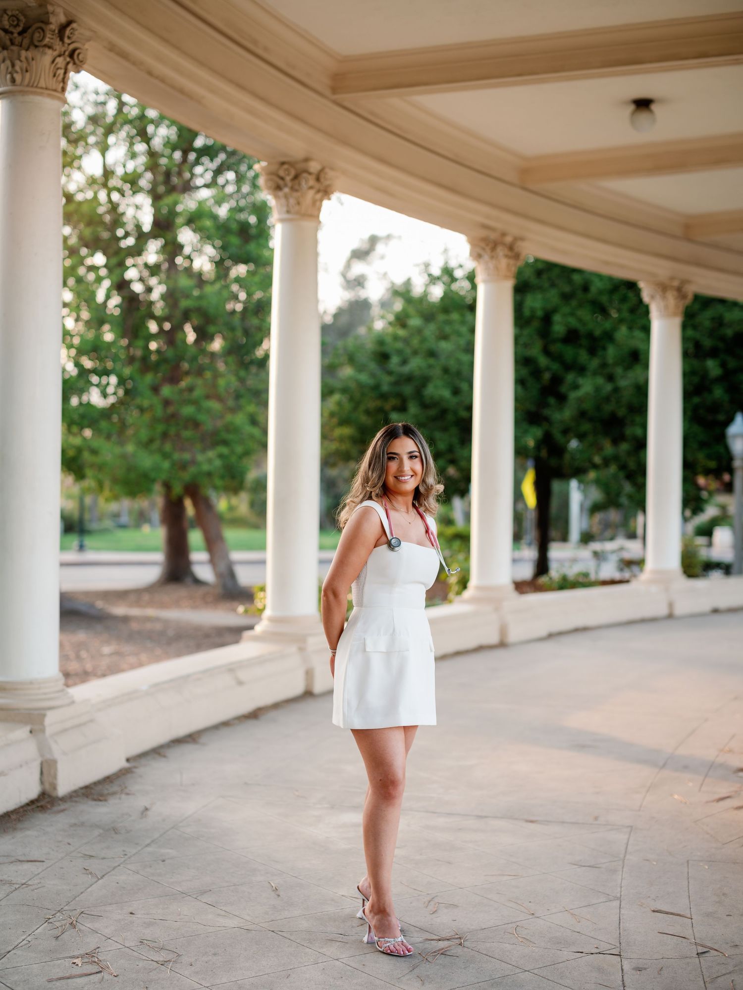 A graduate in white dress poses elegantly under a covered walkway with ornate columns.