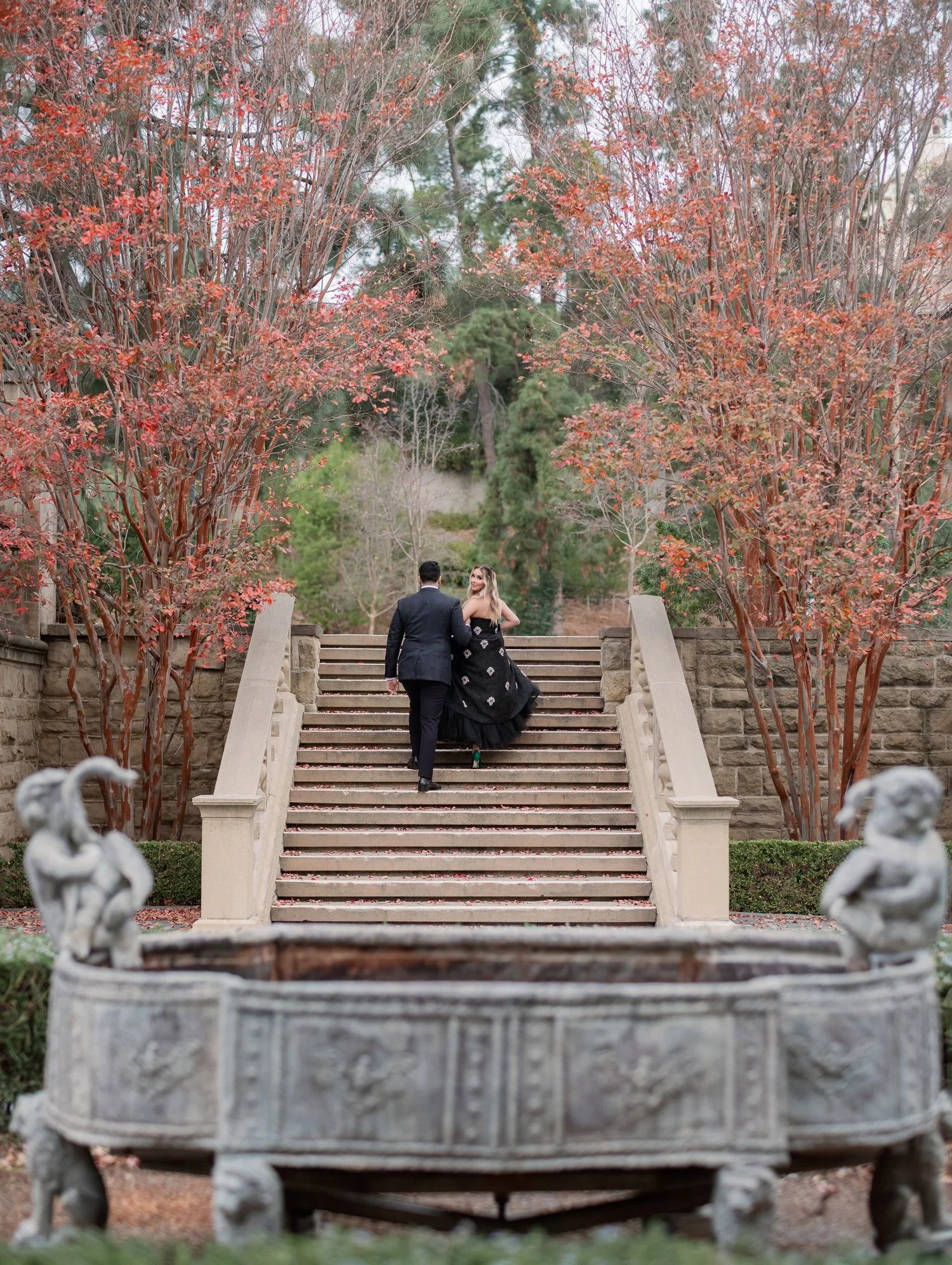 A couple ascending stone steps surrounded by coral-colored trees and ornate stone fountain.