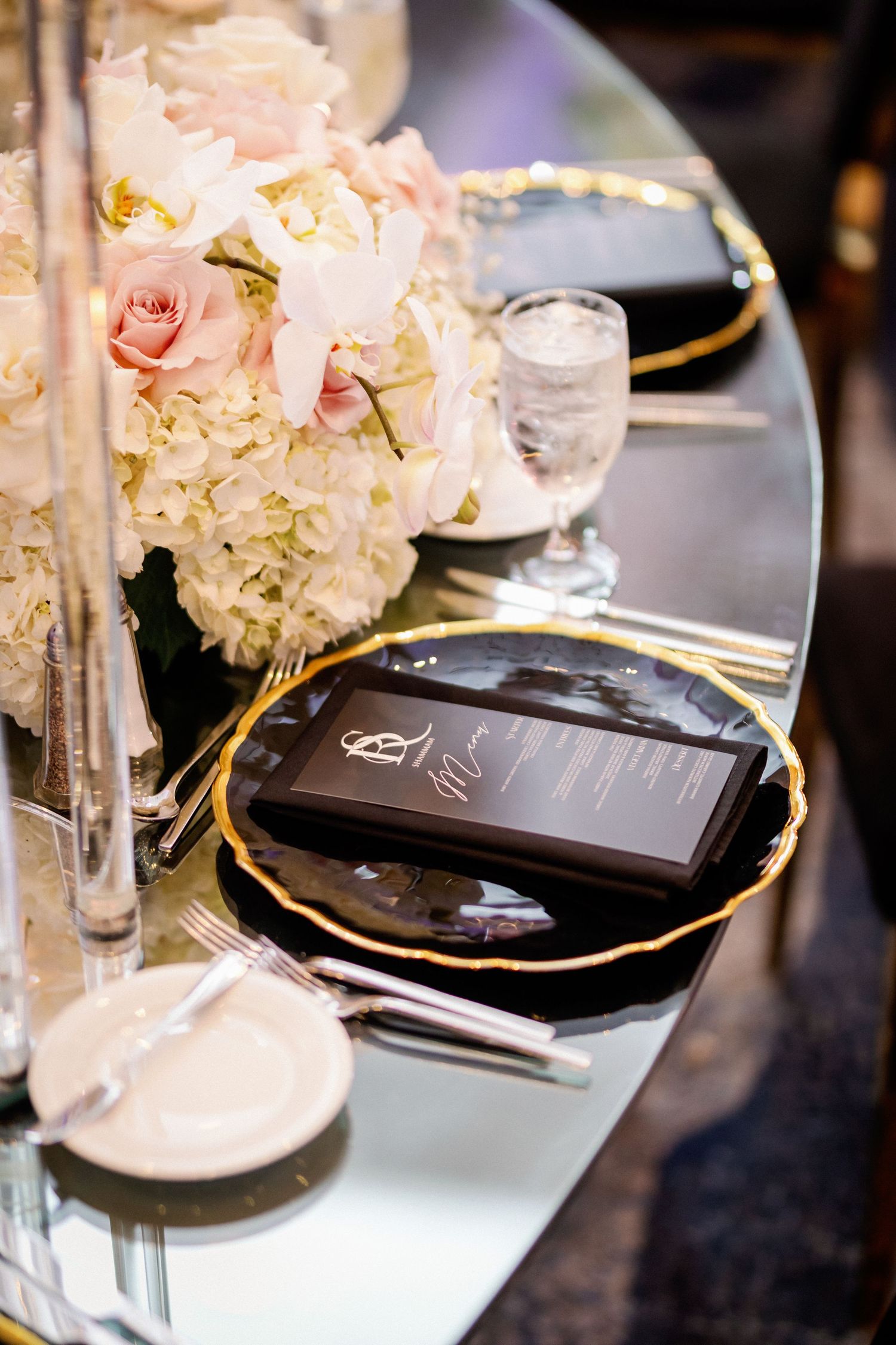 Luxurious table settings with black and gold plates, white hydrangeas, and crystal vases at a formal event.