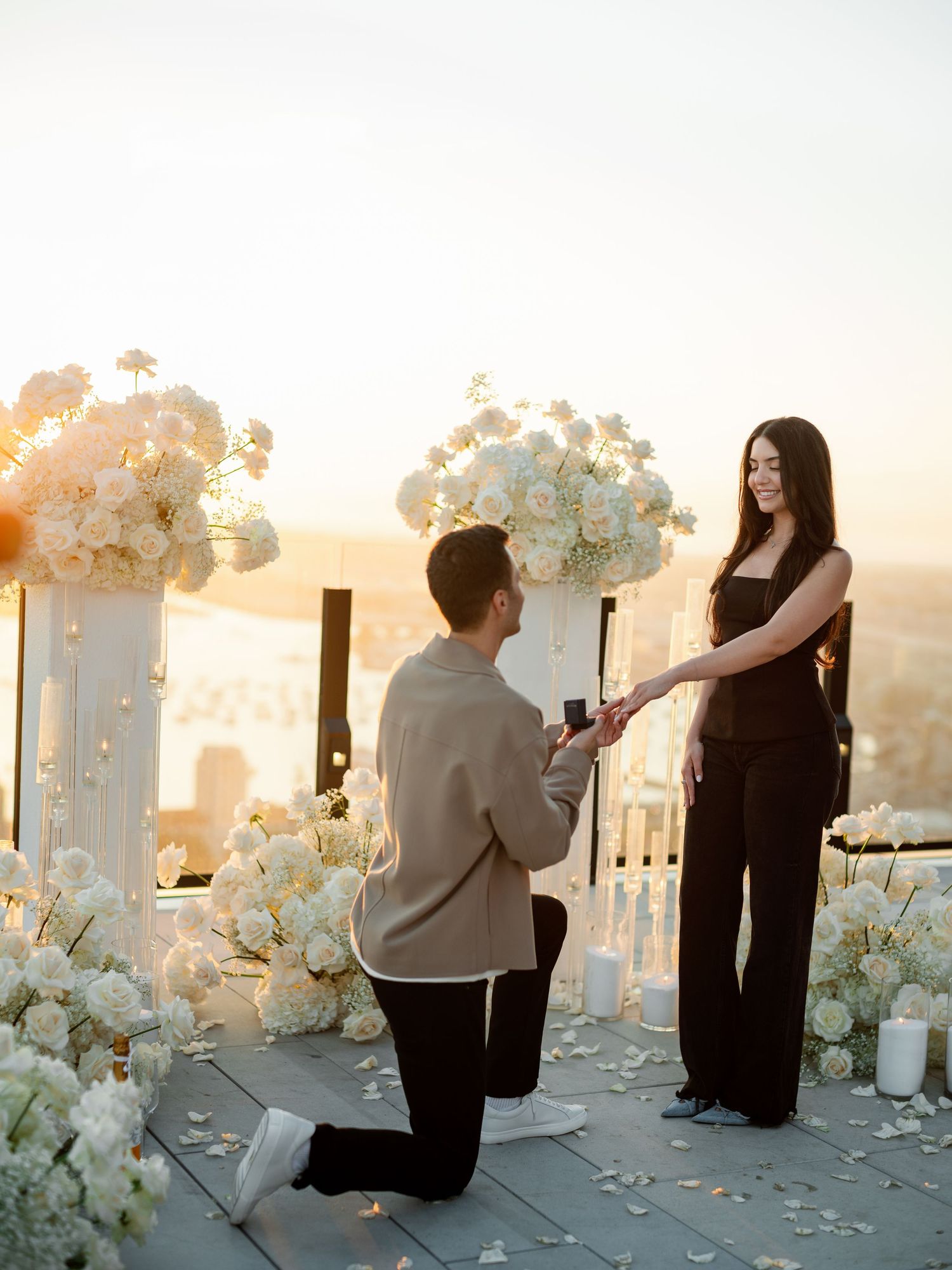 Romantic outdoor proposal setup with white floral arrangements and rose petals at sunset.