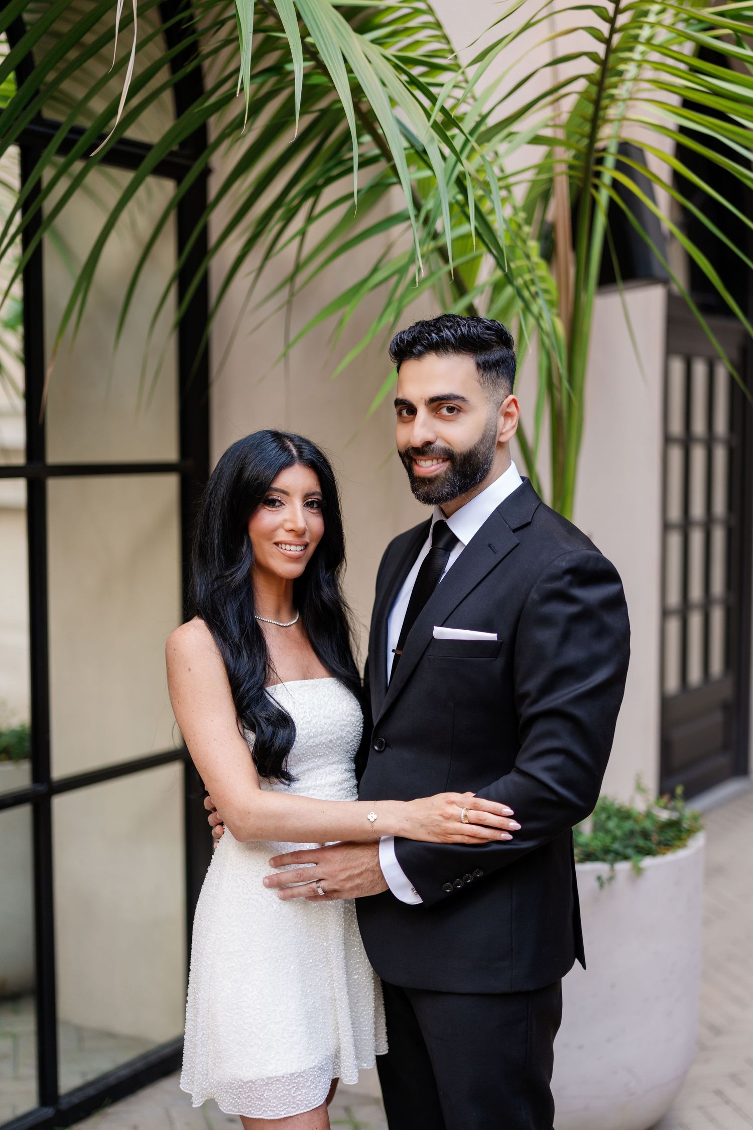 Engagement photos of a couple in formal attire posing against a light colored wall with tropical plants.