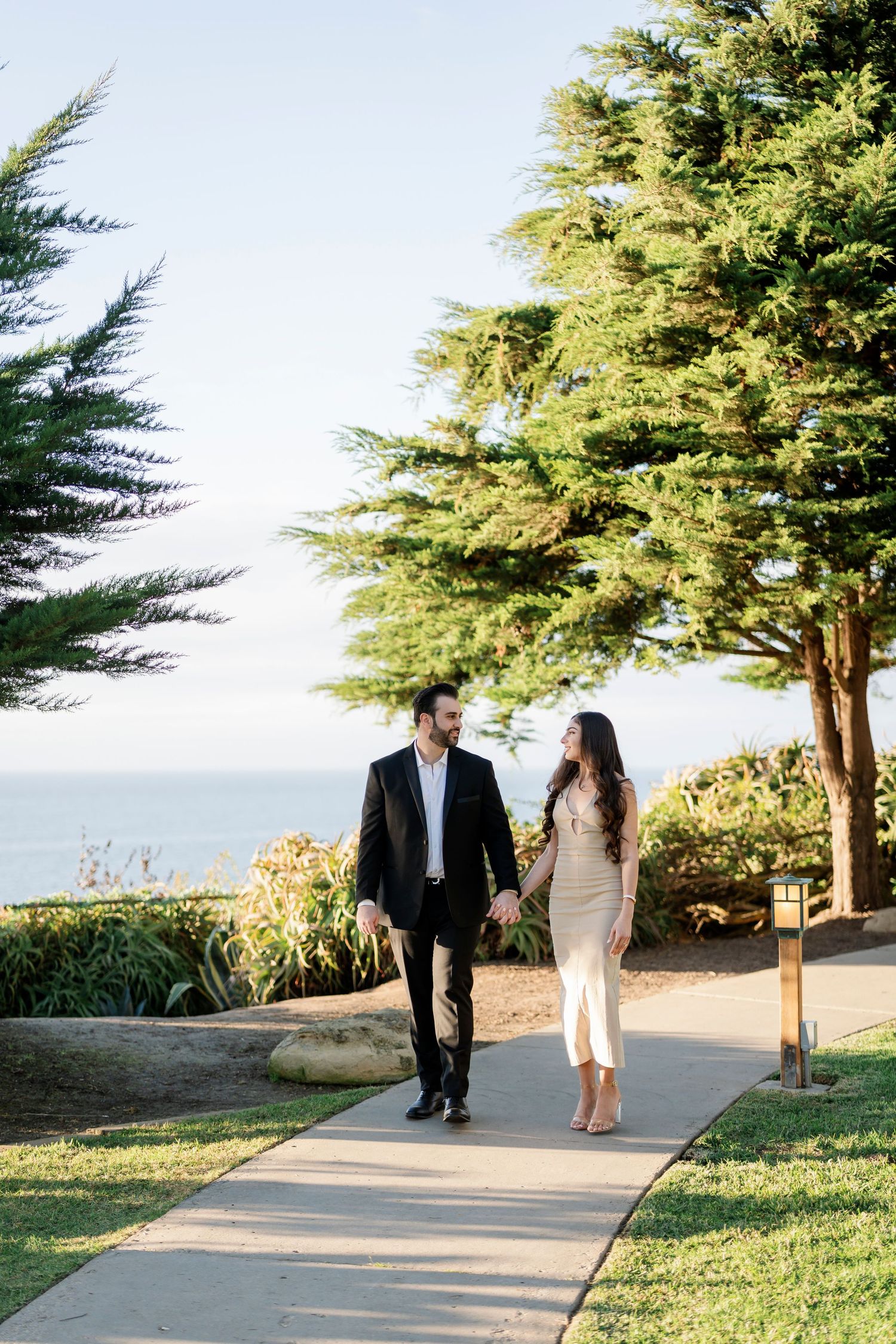 Couple walks together on paved path surrounded by evergreen trees during sunset.