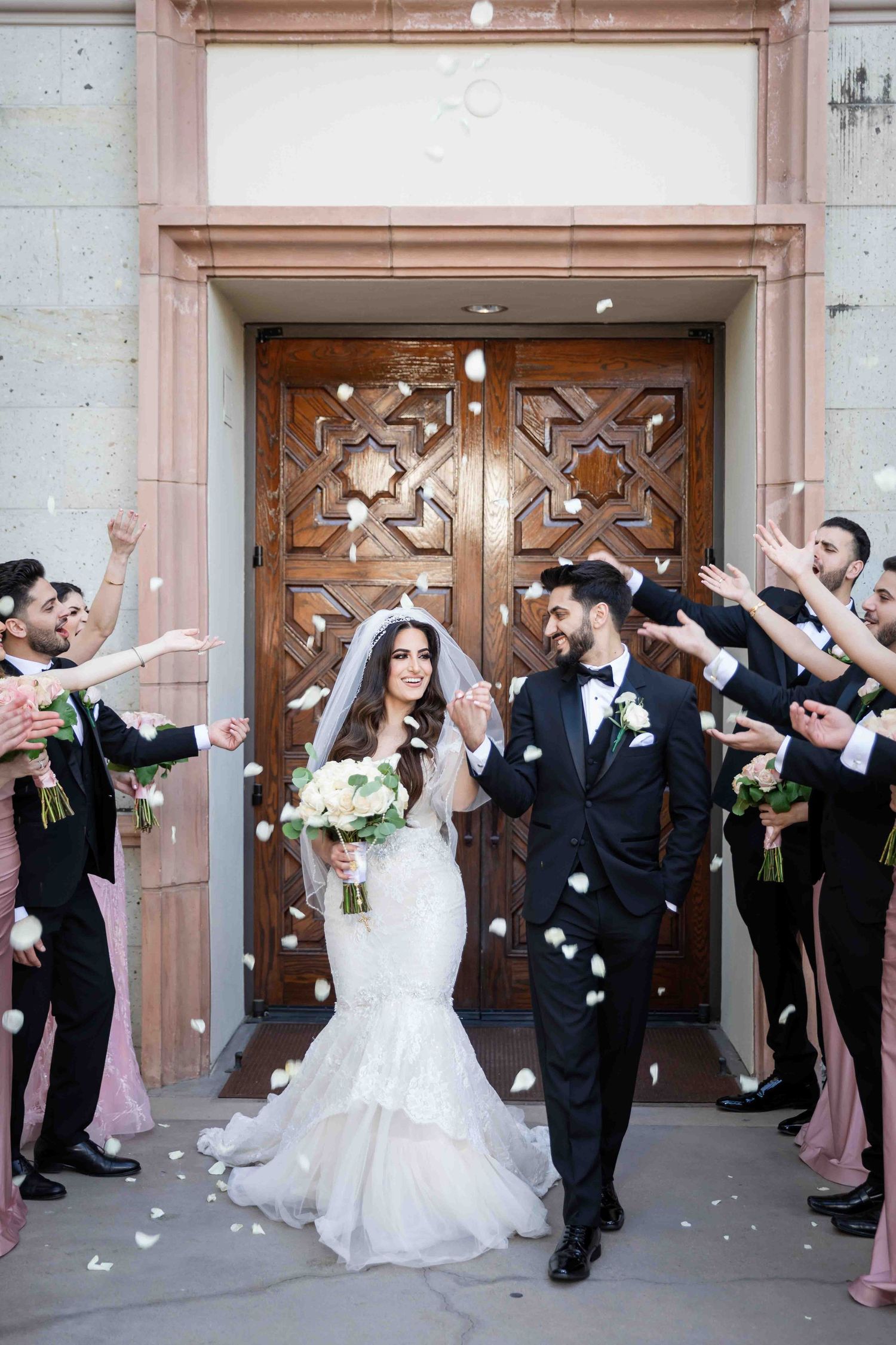 Wedding party celebrates with confetti and joyful poses in front of ornate wooden church doors.