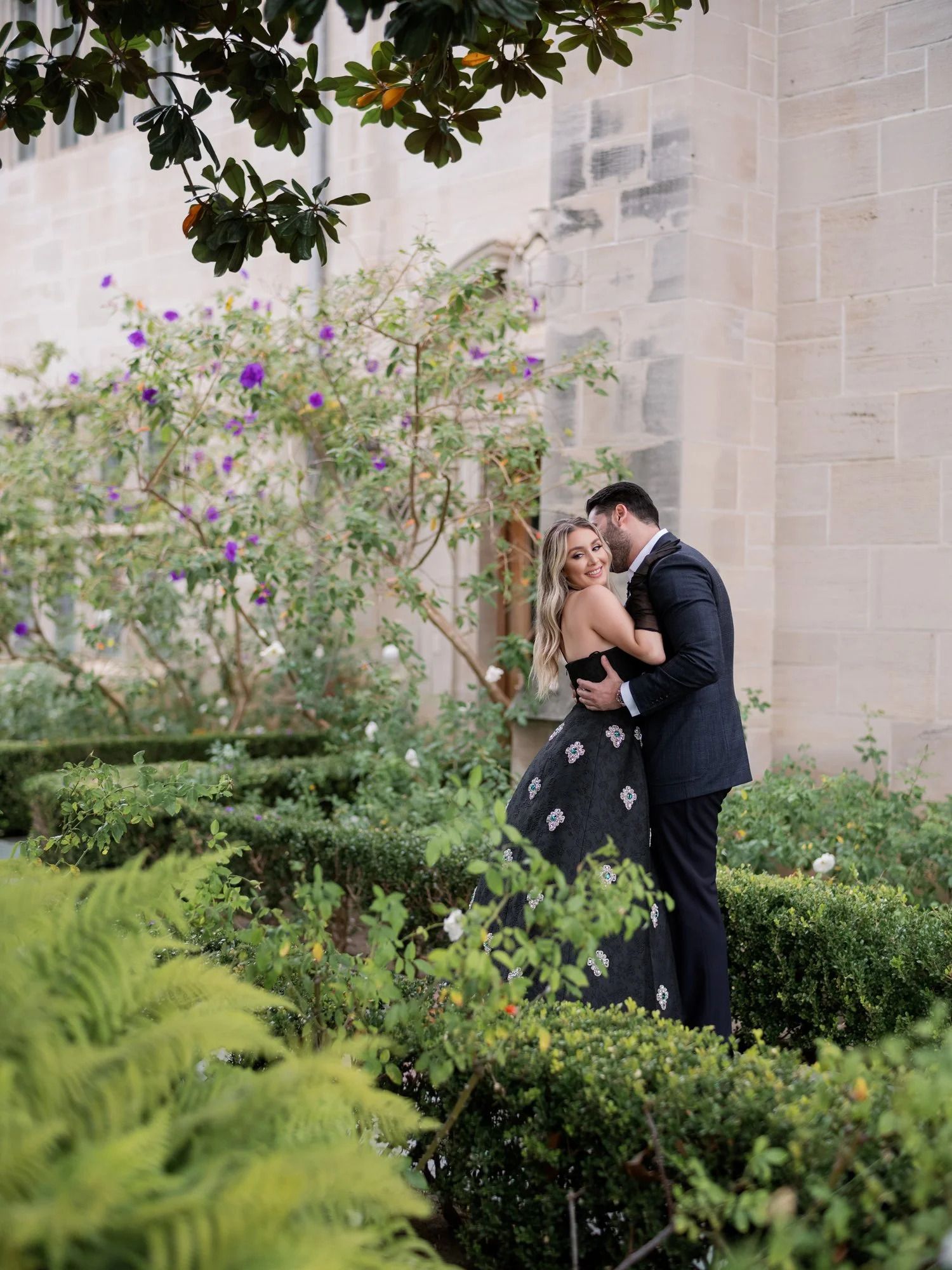 A romantic moment in a lush garden with stone walls and purple flowers.